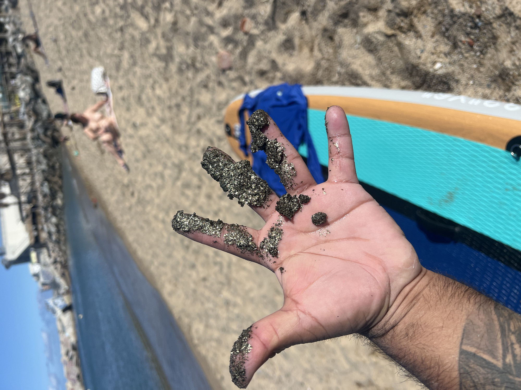 A person holds a hand filled with dark, glittery sand toward the camera. Behind the hand is a blue shirt, a paddleboard, and a beach. The background shows a city on the coast and the Mediterranean Sea. The city is Barcelona, Spain. People are on the beach. The sand is composed of dark, shimmering minerals, adhered to the hand and fingers. The person is likely at Barceloneta Beach in Barcelona.