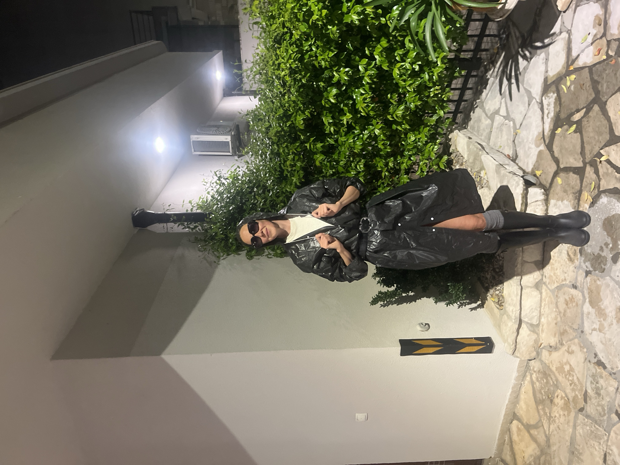 This nighttime image, set in Budva, Montenegro, depicts a person standing outdoors against a backdrop of a building wall and dense green foliage. The subject, positioned slightly to the right of the center, is wearing oversized dark sunglasses, a light-colored top, and a distinctive, shiny black, oversized coat or raincoat that appears to be made of a plastic-like material. They are also wearing dark boots with visible grey socks, and their hands are clasped in front of them.
The background features a light-colored building wall on the left, equipped with a wall-mounted air conditioning unit and a dark drainpipe. Bright, artificial wall lights illuminate the scene, casting strong light and shadows. A lush hedge of green bushes runs along the base of the wall, separating the building from a pathway. The ground is covered with irregularly shaped, light-colored flagstones. Further along the wall to the right, a black sign with two yellow chevron arrows pointing downwards is visible. The person appears calm, posing for the camera. There is no visible text in the image.