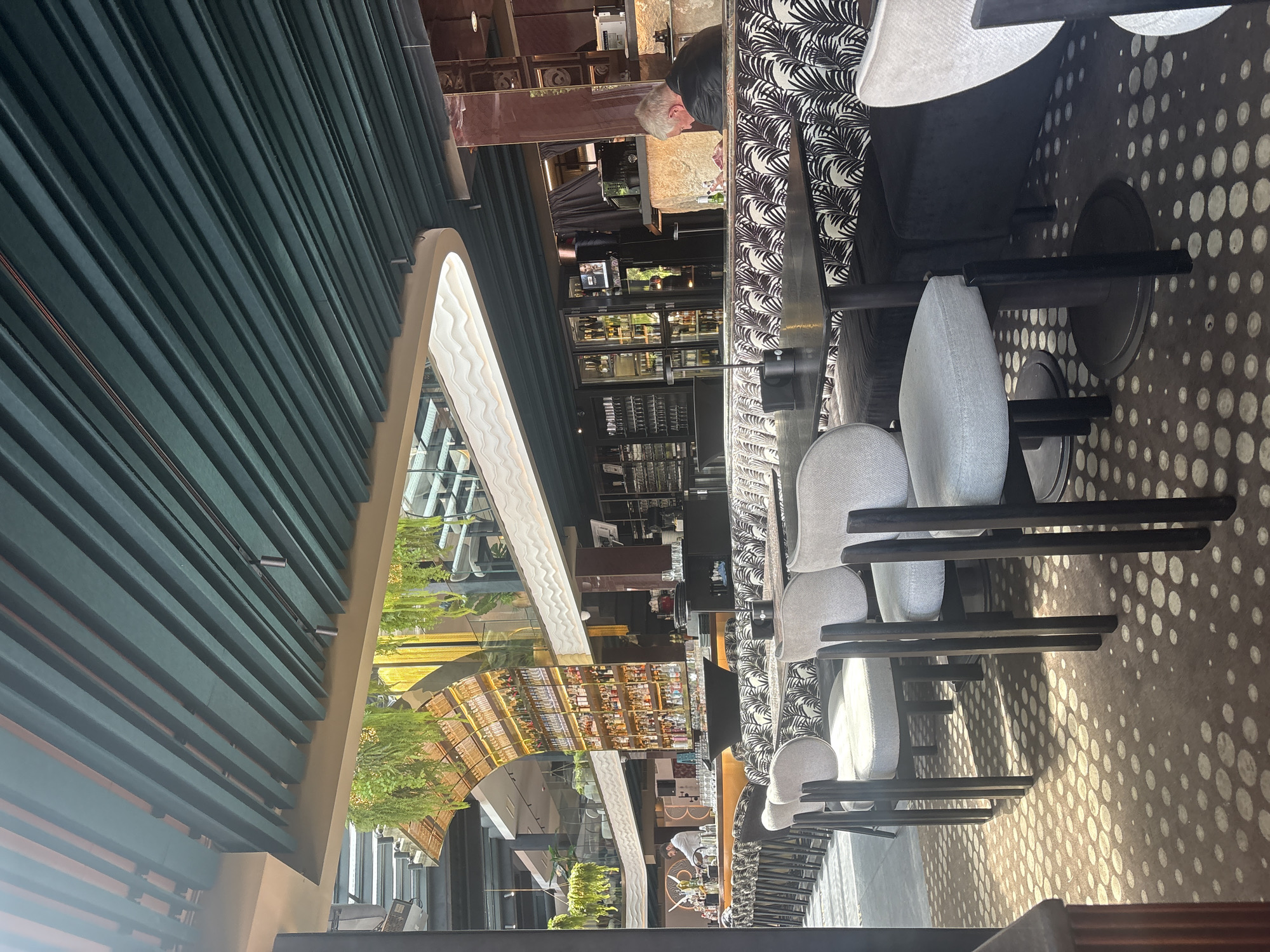 The image shows the interior of a modern, upscale bar or restaurant. The scene is brightly lit, with a focus on the dining area in the foreground and the bar area in the background. The decor features a mix of textures and patterns, including a green slatted ceiling, a white curved light fixture, and seating with a black and white patterned upholstery.
In the foreground, several chairs with light gray cushions and dark wooden legs are arranged around black tables. The floor has a distinctive pattern of light-colored circles on a brown background.
The middle ground reveals a long bar area with high stools and a bartender in a white shirt preparing drinks. Behind the bar is a multi-tiered display of bottles, illuminated by warm lighting. Hanging plants add a touch of greenery to the upper levels of the establishment.
In the background, a man with gray hair is seated at a table, looking down at something in his hands. He is wearing a dark jacket. A bar area with refrigerators and more bottles is visible behind him.
The overall impression is one of a stylish and contemporary establishment designed for socializing and dining. The location is identified as Unknown, Unknown, suggesting the specific city and region are not provided.