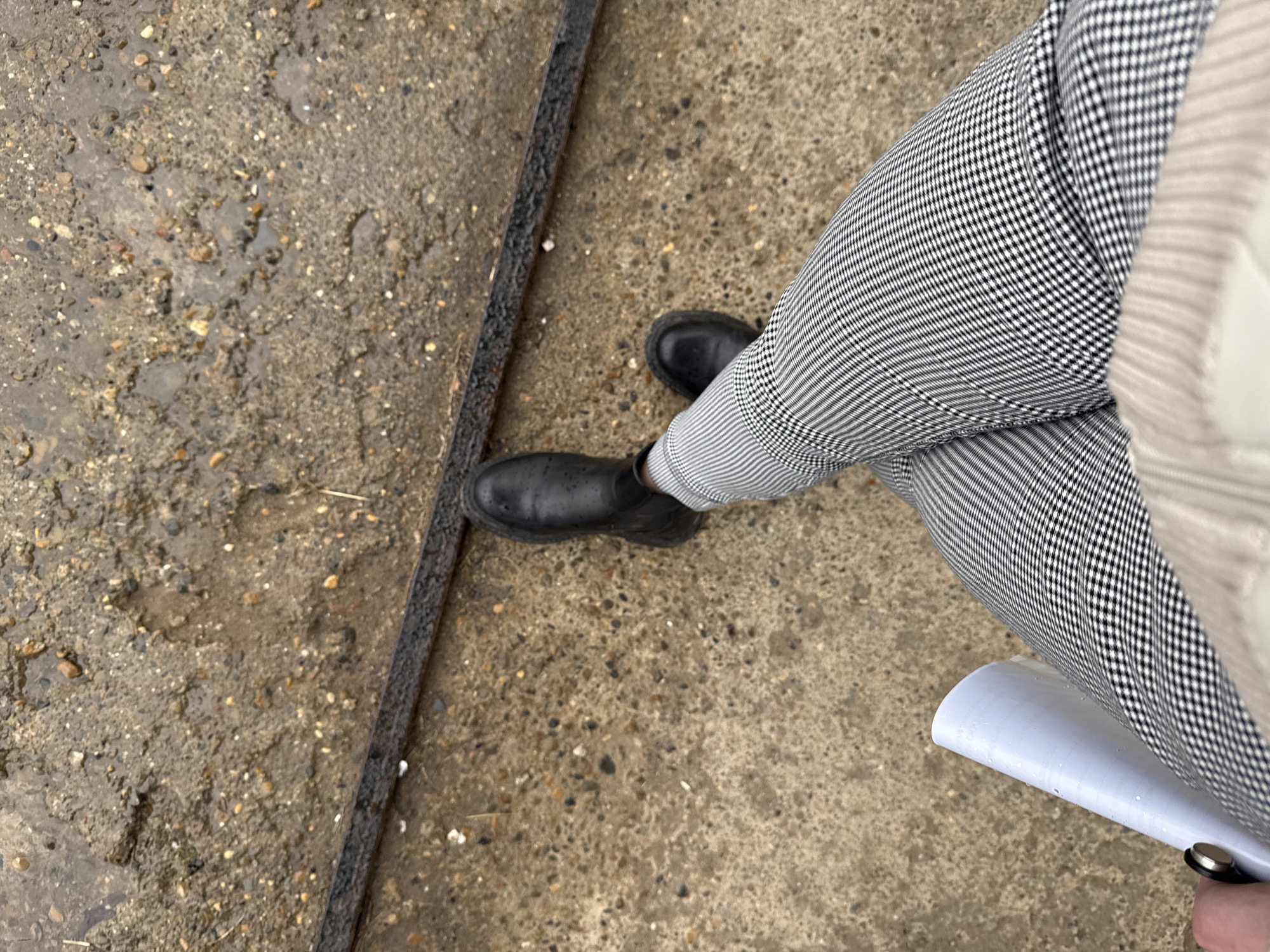 A medium-angle shot depicts a person standing on a wet, mottled concrete surface, likely outdoors. The person is wearing black ankle boots and houndstooth-patterned trousers. A narrow, dark-colored strip, possibly metal or asphalt, lies along the left side of the frame, separating the concrete into two distinct sections. The person is holding a rolled-up white object. The location cannot be verified as being in Chaikovsky, Russia.