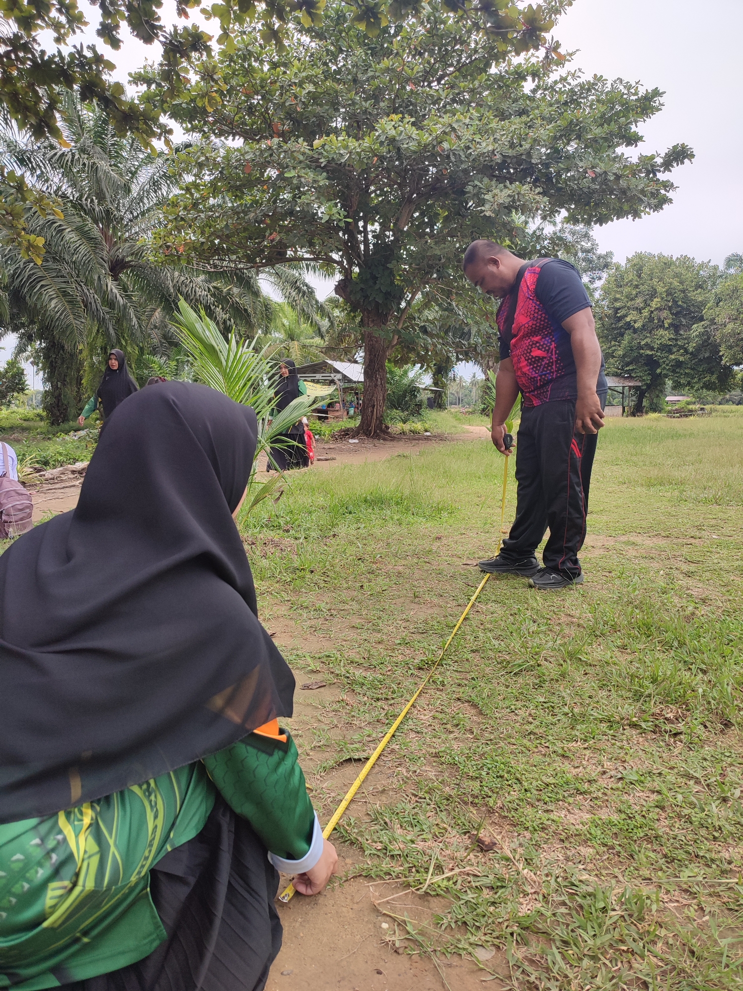 In Blangriek, Indonesia, a man in a black shirt with a pink and red patterned overlay and black track pants is using a yellow measuring tape outdoors. He is bent over, looking down at the tape as it lies on the ground. In the foreground, a person wearing a black hijab and a green long-sleeved shirt with a yellow and green pattern is also looking down at the measuring tape, with their hands near it. Several other individuals, including two women in hijabs, are visible in the background. The scene is set in a grassy area with trees, including palm trees, and what appears to be a small structure in the distance. The sky is overcast.