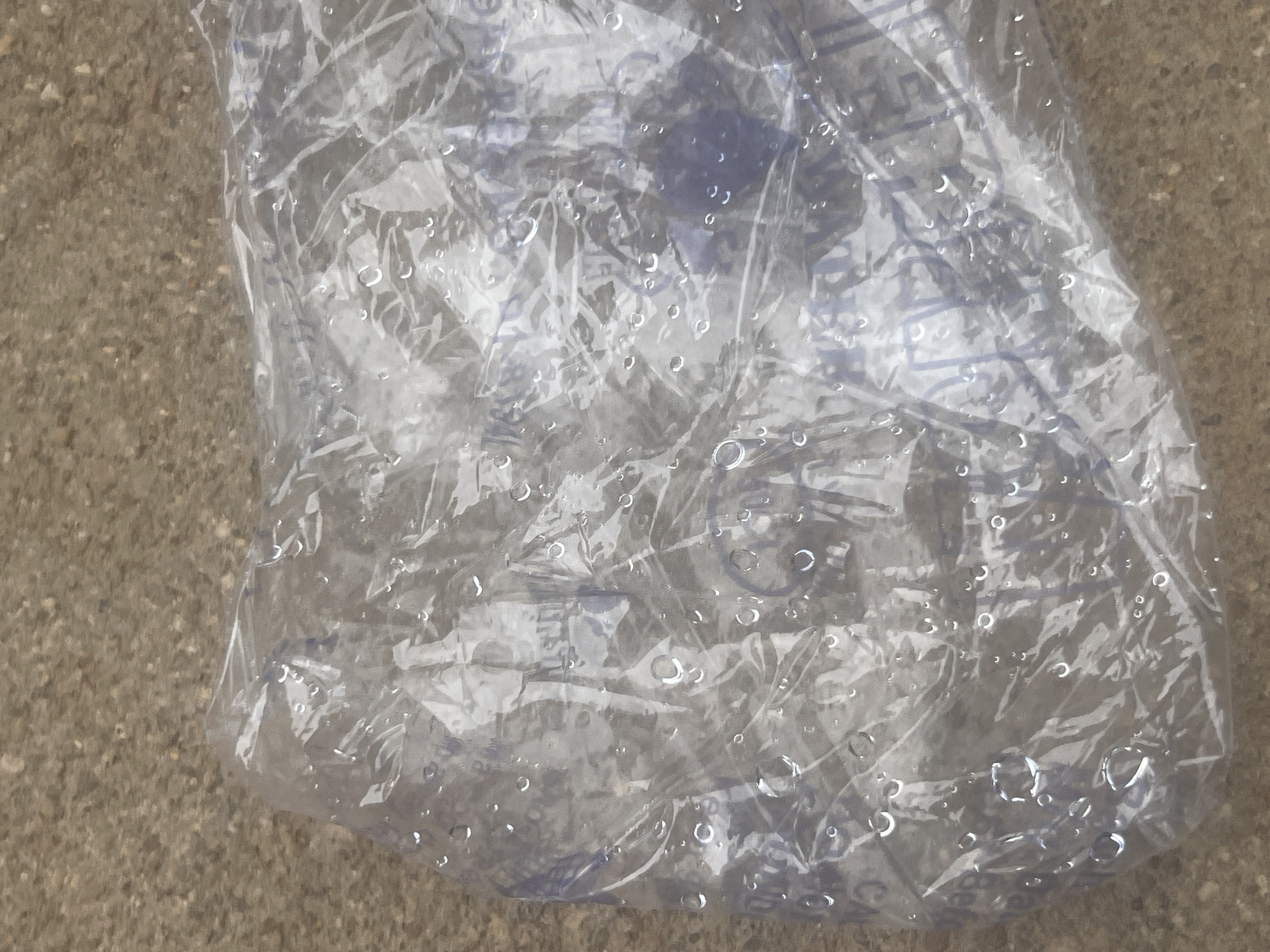 A close-up, overhead shot shows a crumpled clear plastic bag, possibly containing an item, resting on a textured, concrete-like surface. Water droplets are visible on the exterior of the bag, and some faint blue lettering or pattern is visible through the plastic. The scene appears to be outdoors in Jalingo, Nigeria.