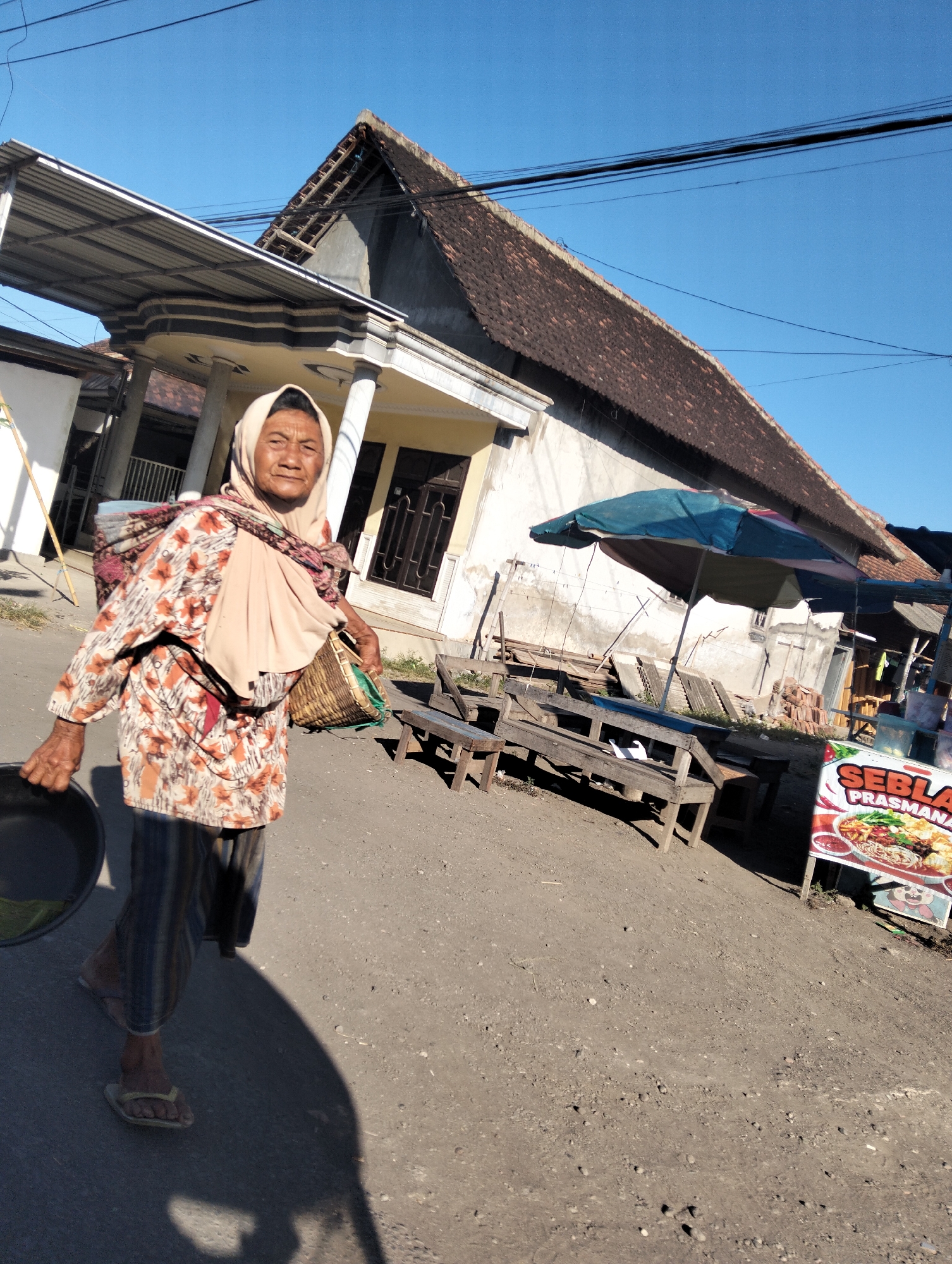 A woman wearing a headscarf and a floral print shirt walks down a dirt road in Kedungringin, Indonesia. She carries a woven basket on her back and a black pot in her left hand. In the background, a house with a tiled roof and a covered porch is visible, along with a blue umbrella and outdoor seating. A sign advertising food can be seen near the seating area. The sky is clear and blue, and the sun casts a long shadow from the woman.