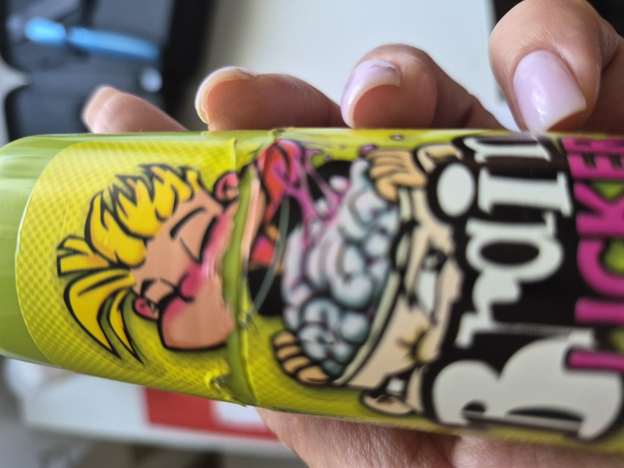 The image shows a close-up of a person's hand holding a bright green cylindrical object, possibly a container or a cosmetic product. The object is wrapped in a colorful label featuring cartoonish characters and text.
The label depicts a cartoon character with blonde hair, large eyes, and a pink shirt. To the right of this character, another figure is visible, seemingly a brain with eyes, surrounded by vibrant colors like pink, white, and black. The overall style is bold and graphic, reminiscent of comic book art.
The text on the label is partially obscured and blurry, but some letters are discernible. We can see "TRO" in large white letters, with a pink letter that appears to be "I" or "L" next to it. Below this, more letters are present, but they are too indistinct to read clearly. The background of the label is a bright yellow with a subtle dot pattern.
The person's hand holding the object has well-maintained fingernails, painted in a light pink color. The fingers are positioned around the cylindrical object, suggesting it is being held or manipulated. The lighting in the image suggests it is daytime, but no specific location cues are apparent beyond the immediate subject. The overall impression is a product with a playful and eye-catching design.
