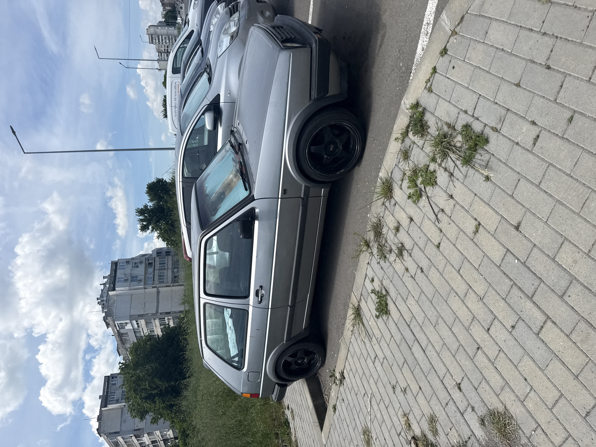 The image shows a silver Volkswagen Golf MK2 parked on a paved sidewalk next to a street in an urban area, likely in Burgas, Bulgaria. The vehicle is positioned close to the curb, with the passenger side facing the viewer. It has black aftermarket rims. There are other parked cars behind it, including a white van with the words "Krasiko Renovation" visible. The sidewalk is made of interlocking gray bricks and has weeds growing in the cracks. Beyond the car, there is a grassy area, trees, and apartment buildings. A street lamp pole is visible in the background against a cloudy sky.