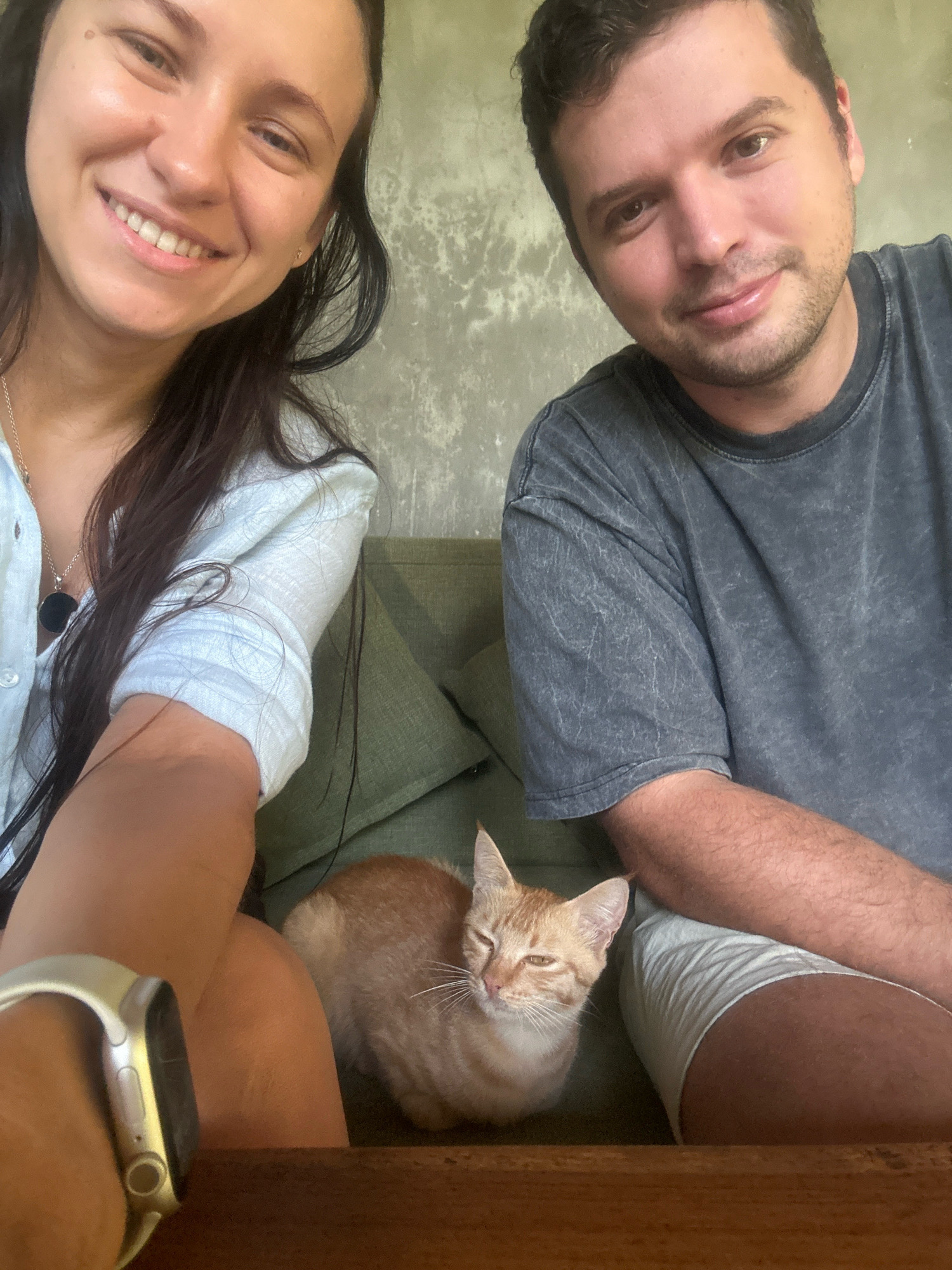 Two individuals, a woman on the left and a man on the right, are positioned in a selfie-style shot. The woman has long, dark hair and is smiling, wearing a light blue collared shirt and a necklace. The man has short brown hair and a beard, wearing a dark gray t-shirt and light-colored shorts. Between them, a ginger cat is lying down, looking towards the camera with its eyes partially closed. The background appears to be an indoor setting with a textured wall and a green sofa. The overall scene suggests a casual, relaxed moment shared between the individuals and their pet. The location is identified as Unknown, Unknown.