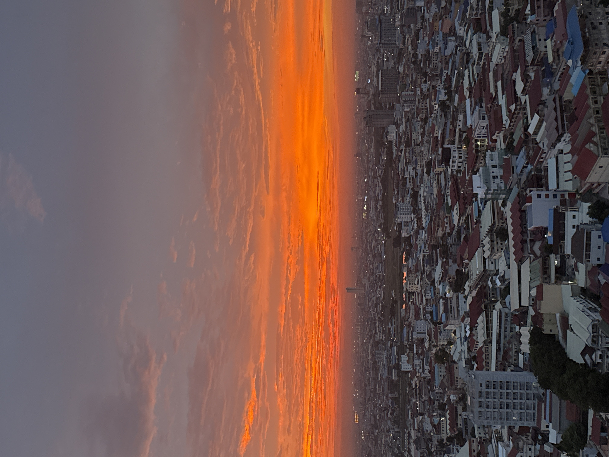 This is a high-angle, vertical shot of a city at dusk, with a vibrant orange and pink sky dominating the upper half of the frame. The sky is filled with streaks of clouds that appear illuminated by the setting sun, transitioning to a muted purple and gray at the top. Below the sky, the cityscape of Khan Mean Chey, Cambodia, unfolds. The buildings are densely packed, with a mix of low-rise structures featuring red and multi-colored tiled roofs, and a few taller buildings, including what appears to be a modern skyscraper in the mid-ground. The scene is devoid of people, and no specific actions or interactions are visible. The environment is urban, characterized by the expanse of buildings and the dusky atmosphere. Lights from some buildings are beginning to illuminate the city.