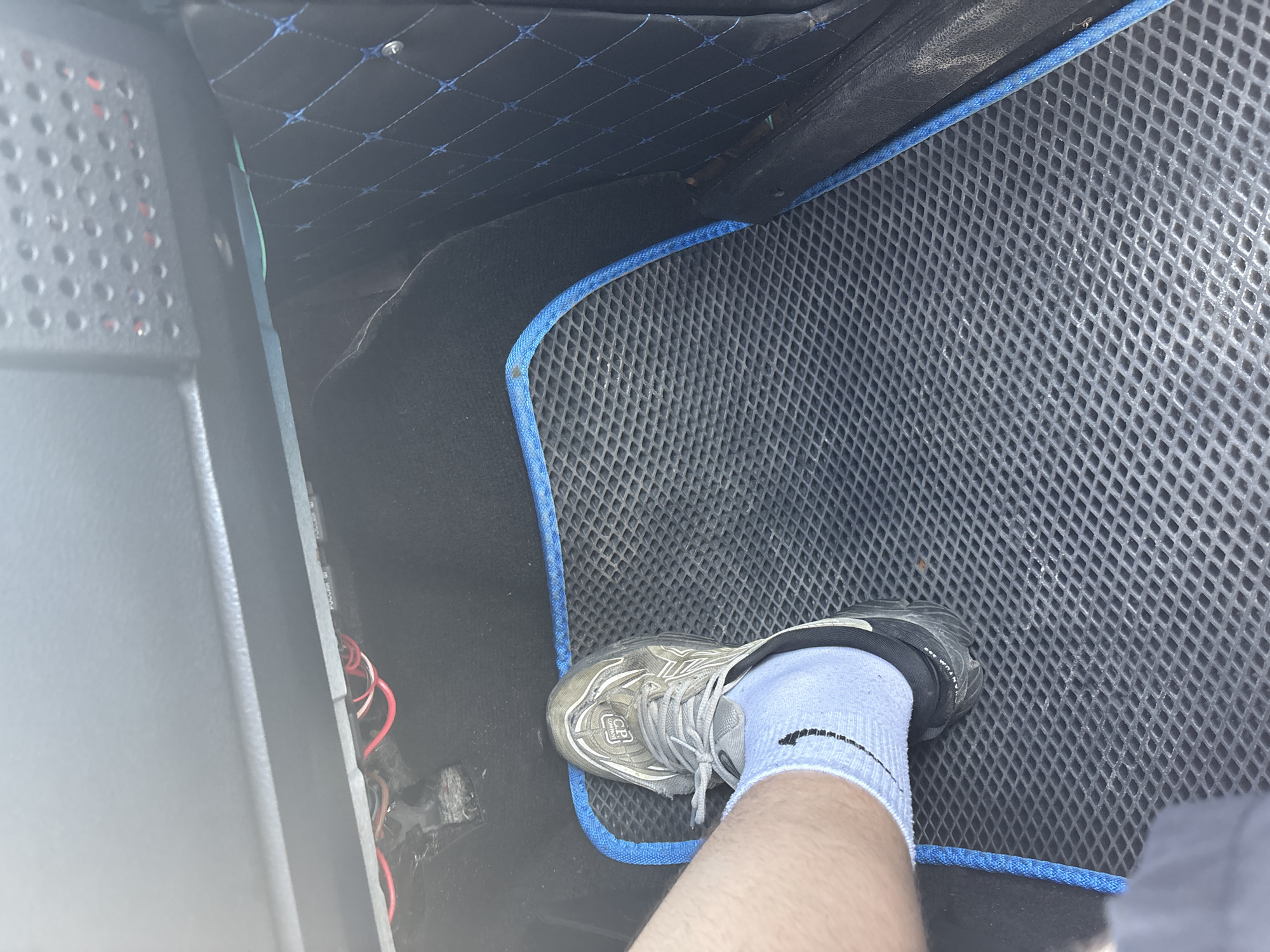 A person's left foot, clad in a light blue sock and a grey athletic shoe, rests on a textured black floor mat with a blue border inside a vehicle. The shoe has a visible logo on its side. To the left of the foot, a portion of a person's leg is visible. Above the mat, exposed red and white wires are visible, suggesting they are part of the vehicle's electrical system. To the right of the mat, a black interior panel with a diamond-pattern stitching in blue is visible. The overall environment appears to be the interior of a car, likely the driver's side footwell.