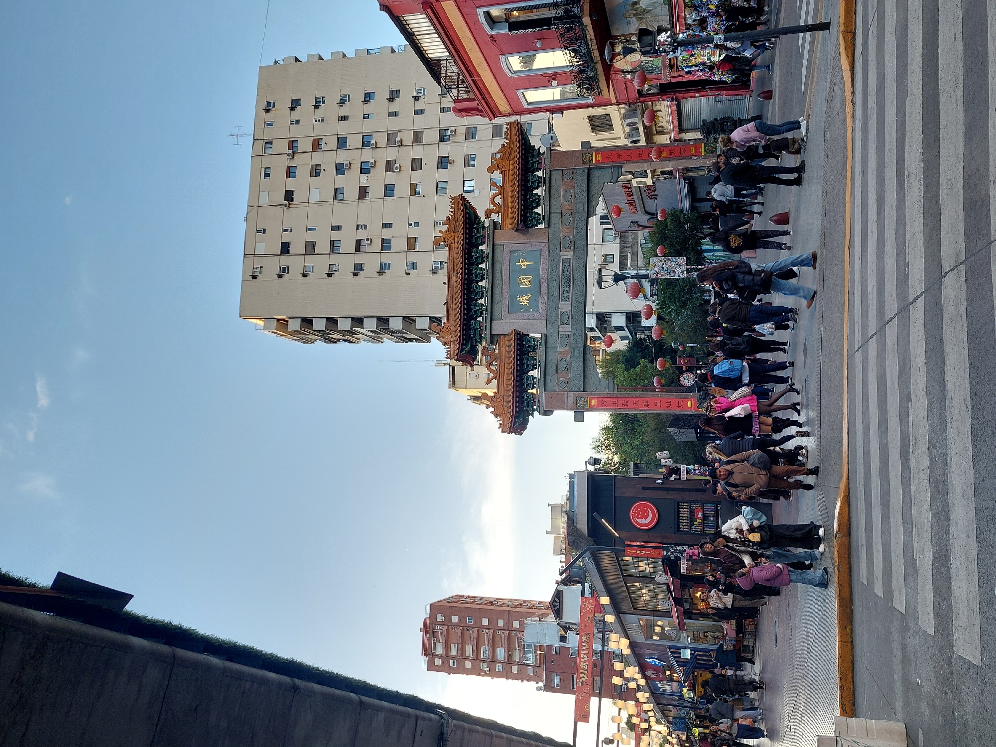The image captures a vibrant street scene in a city, indicative of a Chinatown district, likely in Buenos Aires, Argentina, under a clear, bright blue sky with a few faint clouds. The lighting suggests late afternoon or early evening.
Central to the image is an ornate Chinese archway (Pai-fang), intricately decorated with traditional designs including dragon-like figures on its multi-tiered roof. Golden Chinese characters, prominently displaying "中國城" (Zhōngguóchéng), meaning "Chinatown," are visible on its main beam.
Many people are present, filling the street and sidewalks. A prominent zebra crossing on the right side of the image is occupied by pedestrians waiting or in the process of crossing. Others are seen walking along the street or gathered near storefronts. They are casually dressed, engaged in daily activities.
Buildings line both sides of the street. To the left and behind the archway, a tall, modern high-rise building with numerous windows and visible air conditioning units stands against the sky. Along the street to the right, lower-rise buildings with varied facades, including a distinctive red one, house various shops. Red Chinese lanterns are strung across the street, enhancing the festive atmosphere. Banners with Chinese text, including "中華牌 龍口粉絲" (China Brand Longkou Vermicelli), hang from shops, further establishing the cultural context. A low wall with greenery is visible in the bottom-left foreground.