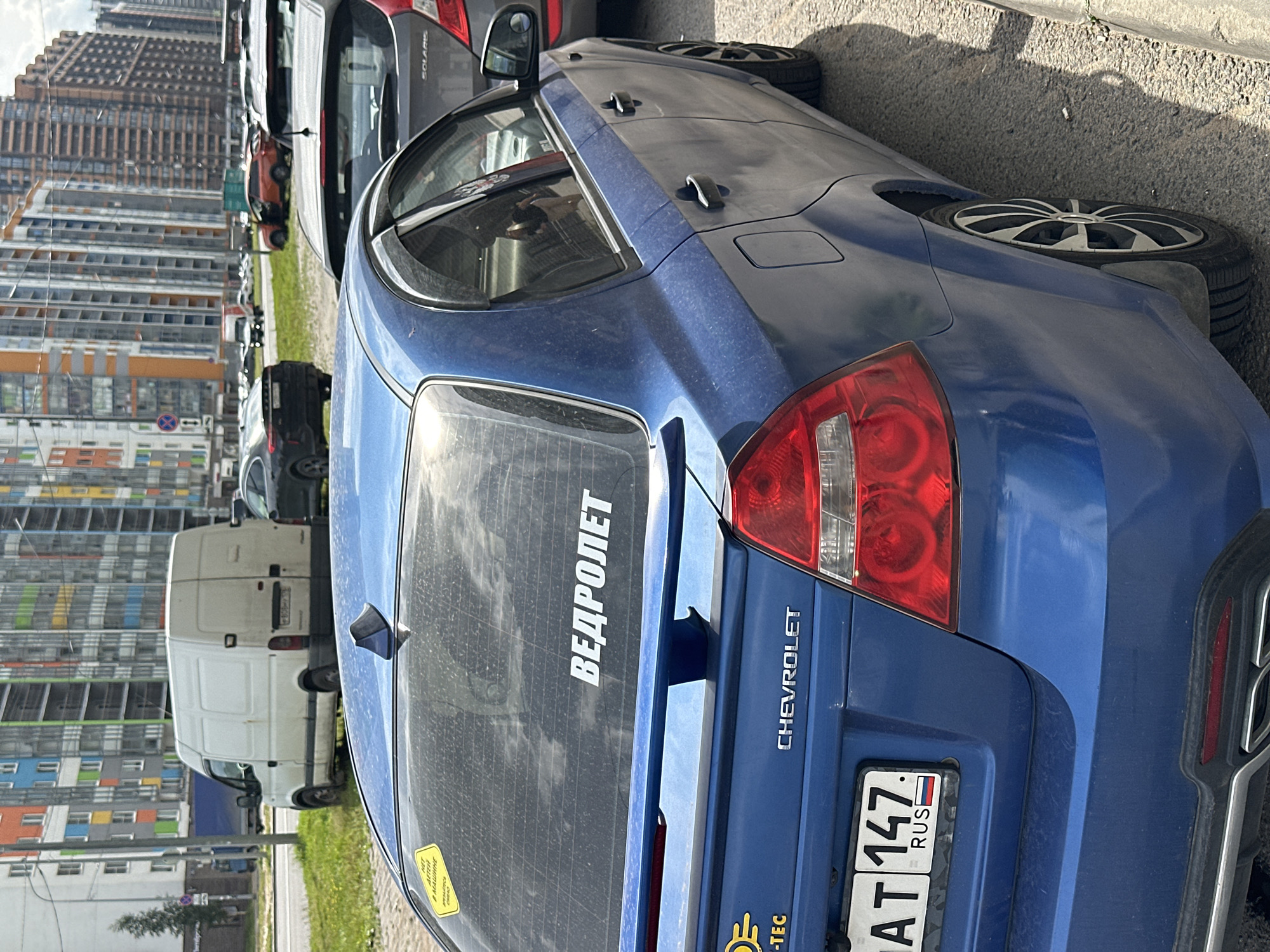 A blue Chevrolet sedan is parked on a street in Murino, Russia. The car has a spoiler on the trunk and a sticker on the rear window that reads "ВЕДРОЛЕТ". The license plate is visible and reads "AT 147 RUS". In the background, there are several multi-story apartment buildings and a white van. Other cars are also parked on the street. The sky is cloudy.