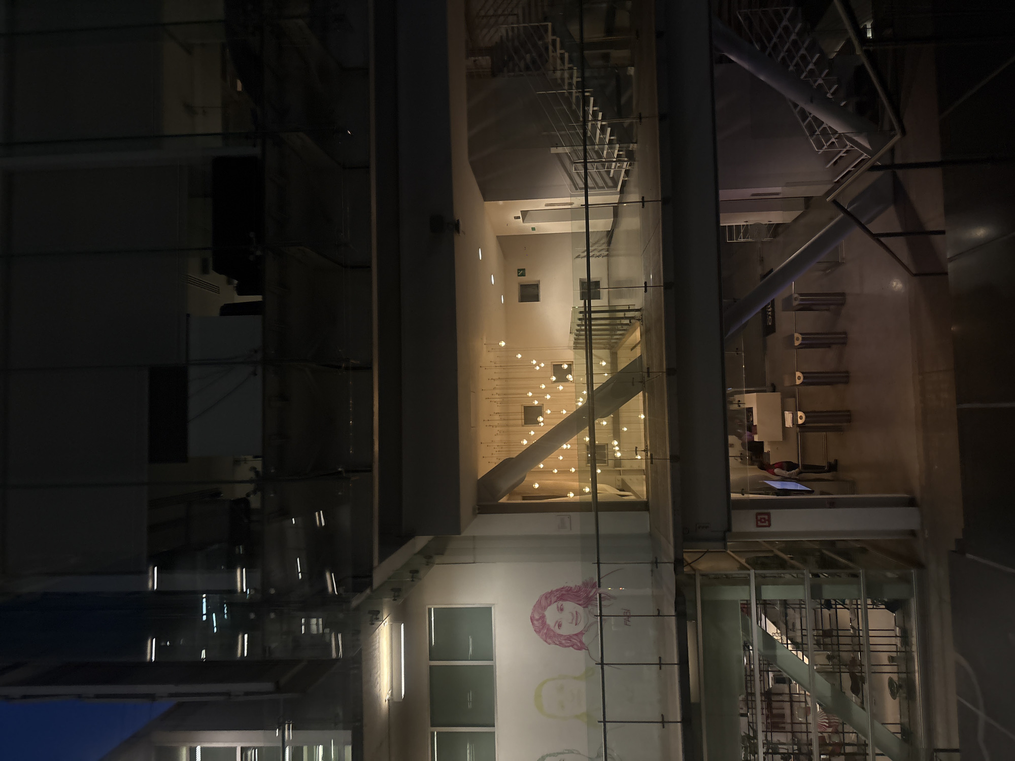 The image displays the interior of a modern building at night, viewed through its glass facade, which also reflects elements from the outside. The building appears to be a multi-story structure with a contemporary design, potentially a public space or a large lobby.
In the upper-middle section, numerous small, spherical lights are suspended from the ceiling at various heights, creating an ethereal, cluster-like illumination. A large, diagonal structural beam cuts across this area.
Below these lights, on a white wall, there is a colorful mural featuring stylized faces, primarily a woman with pink hair. The name "FABI" is subtly visible near this figure. Further down on this wall, rectangular windows are visible.
To the right, metal staircases with glass railings ascend through the levels, showcasing the building's internal architecture. On the ground floor, security turnstiles or access gates are present. Beyond them, a person wearing a red top and dark pants is seated, seemingly looking at a monitor or screen. Another screen is visible on a counter near the turnstiles. The word "metro" is visible on a sign in this area, suggesting proximity to a public transport system.
The lighting is predominantly artificial, originating from the suspended lights and various internal fixtures, creating a warm ambiance. The exterior reflections on the glass facade show dark outlines against a faint dark blue sky. The scene is quiet, indicating a late hour. The location context indicates this is in Mexico City, Mexico.
