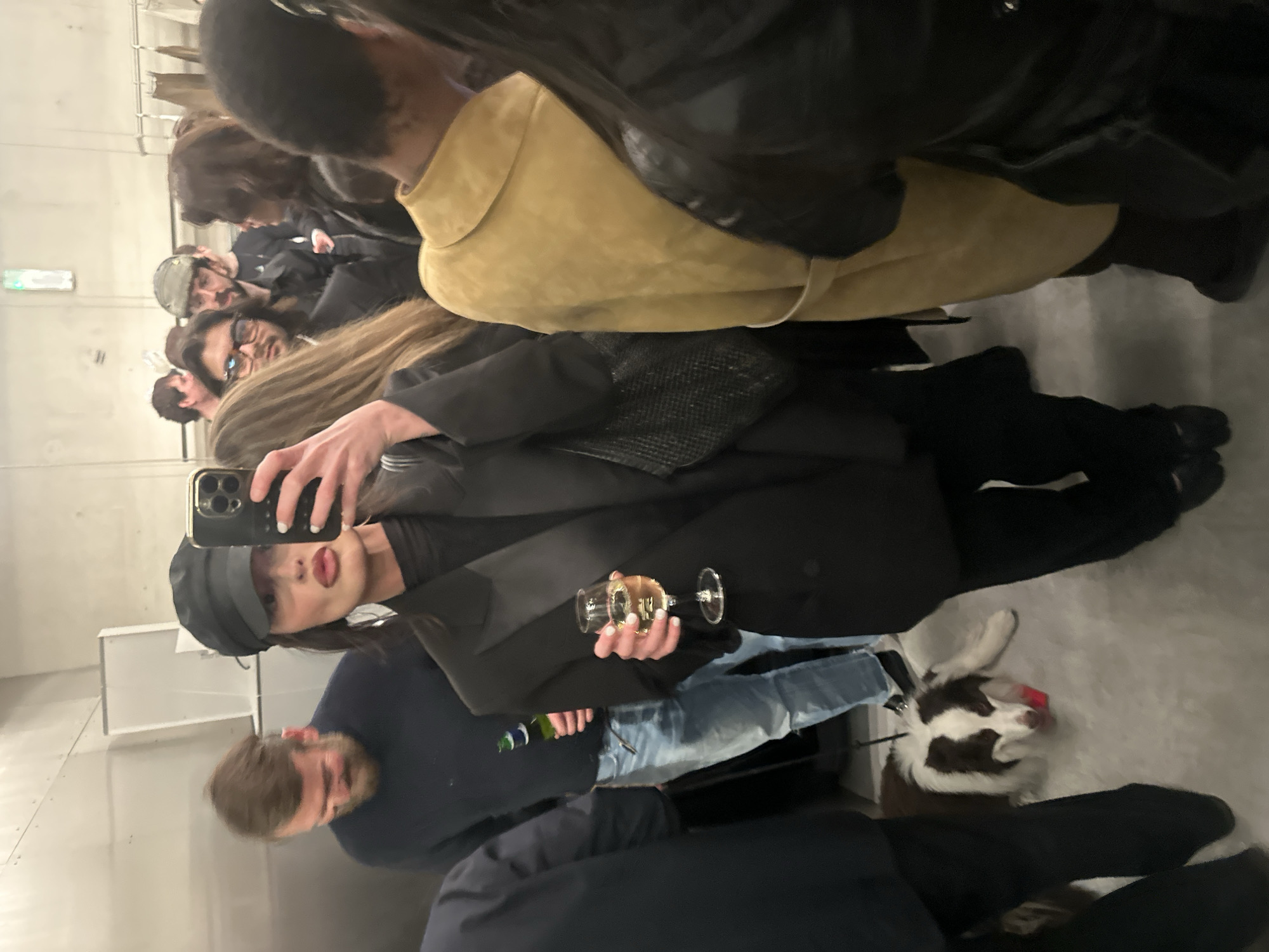 An indoor, crowded scene in a modern commercial or event space in London, United Kingdom. The primary subject is a woman, centered, taking a mirror selfie with a dark-colored iPhone (featuring three prominent rear cameras) held in her right hand. She wears a black baker boy cap, a black blazer over a dark top, and black trousers, holding a wine glass containing a light-colored liquid in her left hand. Her lips are visibly exaggerated, and her fingernails are painted white.
Surrounding her are multiple individuals. To her left, partially obscured, is a man with a beard wearing a dark sweater and blue jeans, holding a green bottle, possibly beer. Below him, a brown and white Border Collie dog lies on a leash on the light-colored floor, a red object near its mouth. Other people are visible in the background and foreground, some looking towards the central figure's phone, others engaged in conversation or standing. One person to the right of the central figure wears a tan or yellow-gold suede jacket. The environment features light-colored walls, possibly metal panels, and a light-colored floor, suggesting a minimalist or industrial aesthetic common in retail or gallery spaces.