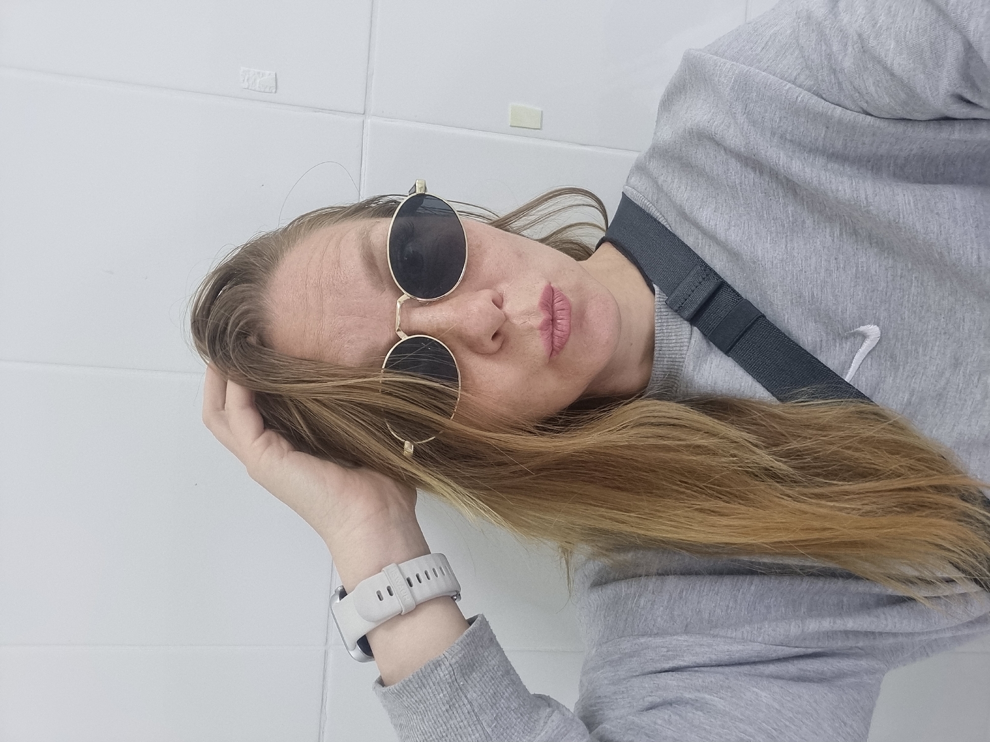 A selfie shows a woman wearing sunglasses and a gray sweatshirt. She has her hand in her hair, and she is looking at the camera with her lips puckered. The woman is wearing a smartwatch on her left wrist. A gray strap from a bag is visible across her chest. The background is a white tiled wall. The image is taken in the city of Chaikovsky, Russia.