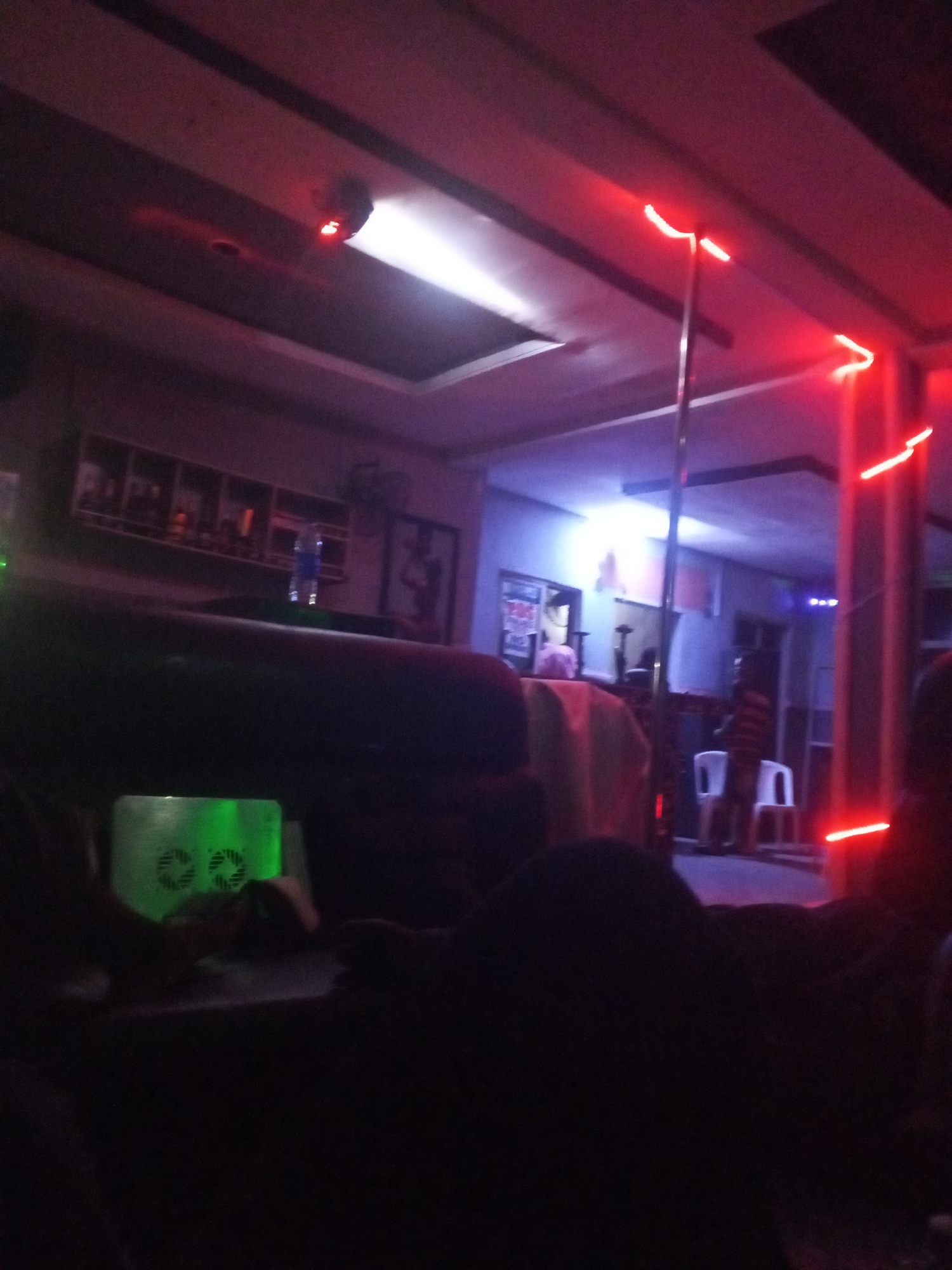 This is a dimly lit indoor scene, likely a bar, club, or lounge, characterized by atmospheric lighting and a social setting.
In the foreground, dark silhouettes of people occupy lounge-style seating, with their forms largely obscured by shadow. On a table in front of them, a rectangular, green glowing device with two circular vents is visible.
The midground features a prominent metallic pole, resembling a stripper pole, positioned towards the right of the frame. Red LED strip lights illuminate the ceiling and a vertical pillar to the right, creating a vibrant glow. To the left, a bar-like shelf unit holds several bottles and other items, with framed pictures or posters on the wall behind it. A white ceiling panel with a bright white light fixture contrasts with the darker surroundings. Another red light source, possibly a smoke detector or alarm, is visible on the ceiling.
Further in the background, several indistinct figures are visible, some standing near the pole, and two white chairs are placed against a wall. More red strip lights are visible on the ceiling and walls in the deeper background. The overall ambiance suggests a nocturnal environment focused on entertainment and socializing. No discernible text is clearly visible.
