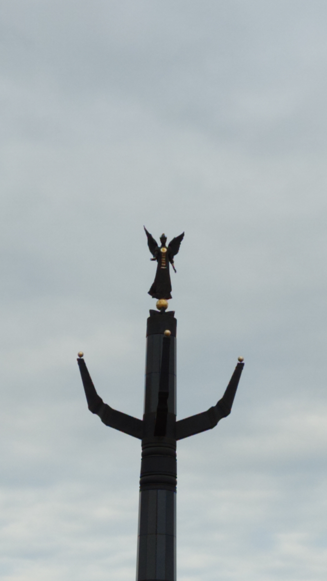 A statue of a winged figure, likely an angel, stands atop a tall, dark monument against a hazy, overcast sky. The statue is silhouetted and appears to be dark, possibly bronze or iron, with outstretched wings and a long robe. It is positioned on a spherical base, which sits atop a multi-faceted, dark gray column. The column has horizontal bands and is topped with what appears to be a stylized crown or a series of protrusions. Two smaller, golden spheres are visible on the monument's structure, one on the main column and another on a separate, pointed element to the left. The overall impression is that of a monument located in the city of Mirny, Russia, possibly a memorial or a landmark.