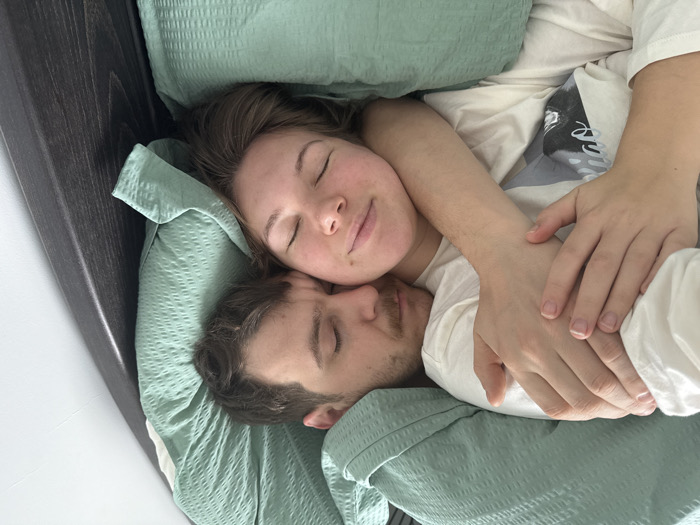 Two individuals, an adult male and an adult female, are depicted asleep or resting embraced on a bed. The male, with short brown hair and light facial hair, wears a white t-shirt. The female, with brown hair, wears a white top that shows a partial dark graphic design. Both have closed eyes. They are covered by a light green, textured duvet or blanket. The male's right arm is positioned over the female's left arm, with her left hand resting on his arm. A dark brown wooden bed frame is visible on the right, against a plain white wall. The indoor scene is brightly lit. This scene is located in Vozdovac Urban Municipality, Serbia.