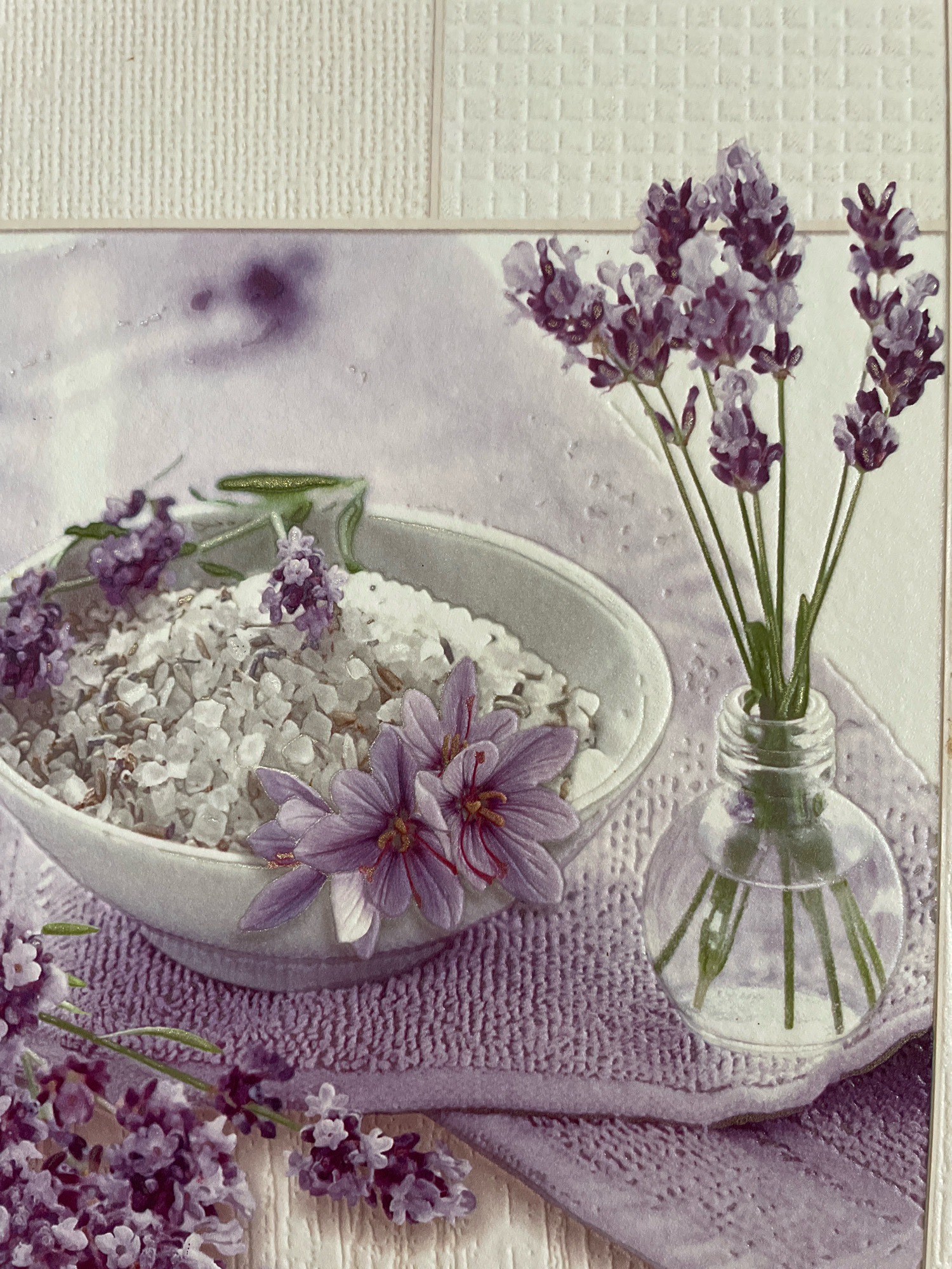 The image depicts a still life arrangement of lavender flowers, bath salts, and a small glass vase, likely intended to evoke a spa or relaxation theme. The scene is set against a textured, light-colored background that appears to be a decorative paper or canvas.
In the foreground and midground, a white ceramic bowl is filled with coarse white bath salts. Several sprigs of lavender are arranged around the bowl and within it. Two larger purple flowers, possibly irises or a similar bloom, are placed on the edge of the bowl, with their stamens visible. To the right of the bowl, a small, clear glass bottle with a rounded base contains several stems of lavender. The flowers are a deep purple and lavender color, with some lighter purple and white accents.
The overall color palette is dominated by shades of purple, white, and light gray, creating a soft and calming atmosphere. The textures in the image suggest a tactile experience, with the granular bath salts, the delicate petals of the flowers, and the ribbed texture of a purple fabric or towel beneath the bowl. The lighting is soft and diffused, casting subtle shadows that enhance the dimensionality of the objects. There are no people or animals present in the image. The scene is static and appears to be a staged photograph or artwork. The location is unknown.