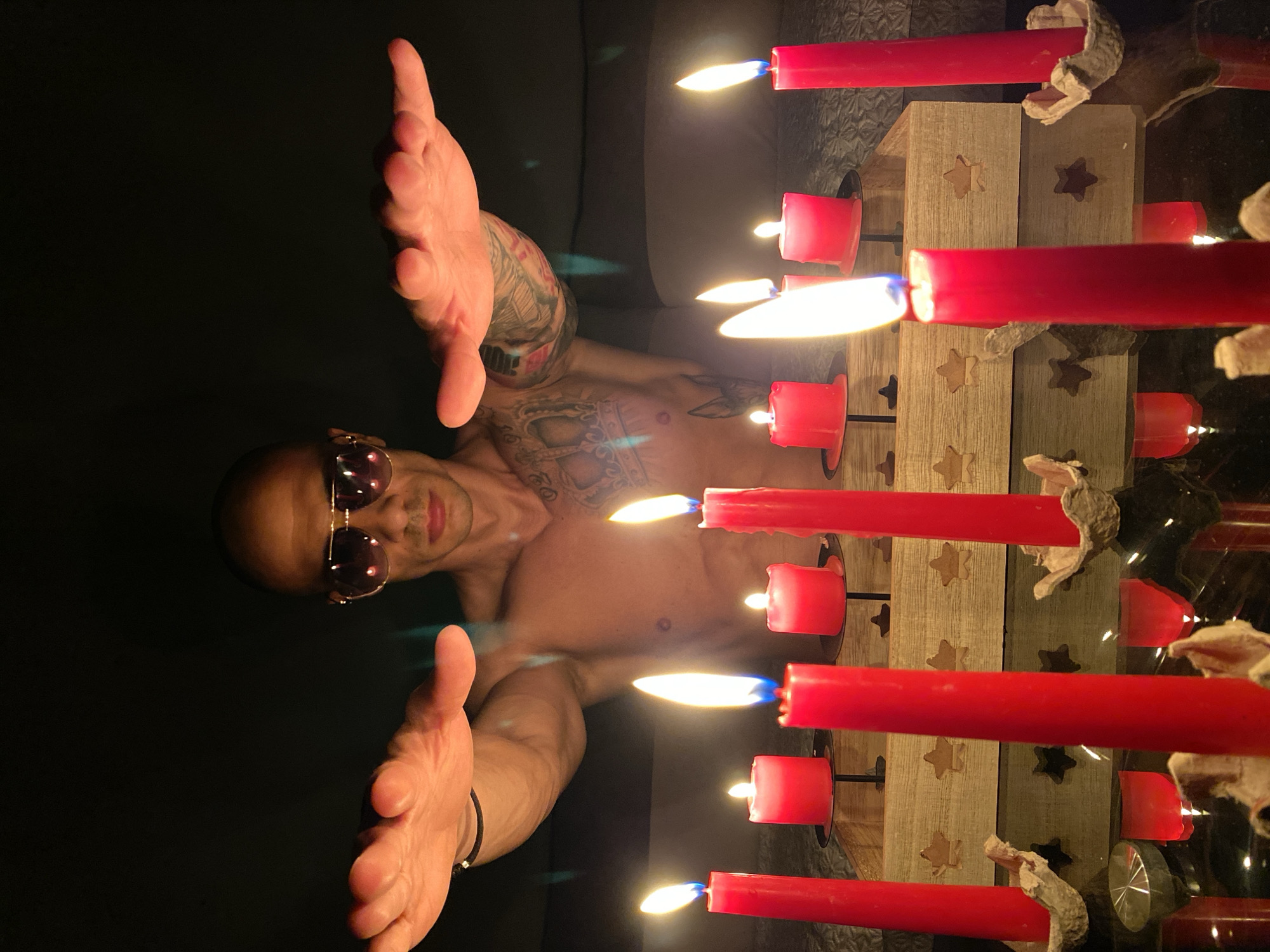 The image captures a dramatic scene in a dark, likely indoor setting, primarily illuminated by candlelight. A shirtless man with a shaved head and sunglasses lies horizontally in the frame, extending both hands towards the viewer with palms open. He is heavily tattooed on his arms and chest, with various intricate designs visible.
To the man's right, a line of red candles stands upright, their flames casting warm light. Some candles are held in simple, crumpled-looking foil or paper holders, while others are set into a rustic wooden candelabra featuring carved star-shaped cutouts. The lit candles and their fiery glow are clearly reflected on a dark, glossy surface beneath them, indicating a mirror or a highly polished table.
The background is almost entirely black, emphasizing the subjects and the light from the candles. The time of day appears to be night or a very dimly lit room, with the candles serving as the main light source. The man looks directly into the camera, his expression partially obscured by sunglasses, suggesting a deliberate pose. No discernible text is visible in the image. The location is contextualized as Pleven, Bulgaria.