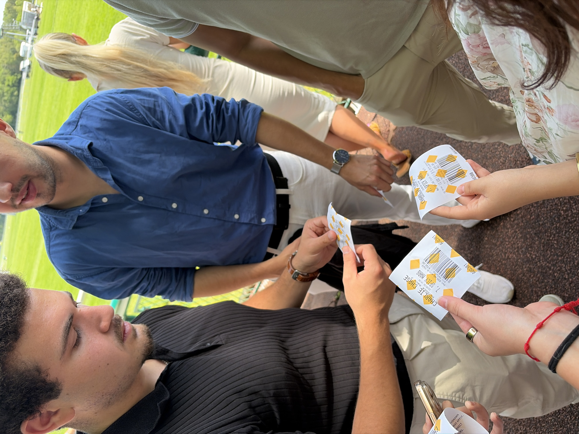 A daytime scene captures a group of individuals outdoors, potentially at a racetrack based on the presence of betting slips. The environment is suggested by green grass in the background, and what appears to be parked vehicles in the distance. The perspective is from above, looking down at the group. Several individuals are visible, each dressed in casual attire. One man wears a black ribbed polo shirt, beige pants, and holds a betting slip. Another man wears a blue button-down shirt and white pants. A woman with long blonde hair stands behind them. Another individual has on a floral patterned shirt and beige pants. The focus of the image seems to be on the betting slips held by the people. The slips have identical information regarding a horse race named "CHARLIE CHASE" and "SPICY MARG" is one of the selections. The slips also denote that the bet was placed on May 23, 2026, at 14:58 for £20.00, with a potential win of £150.00. The scene suggests a moment of shared activity.