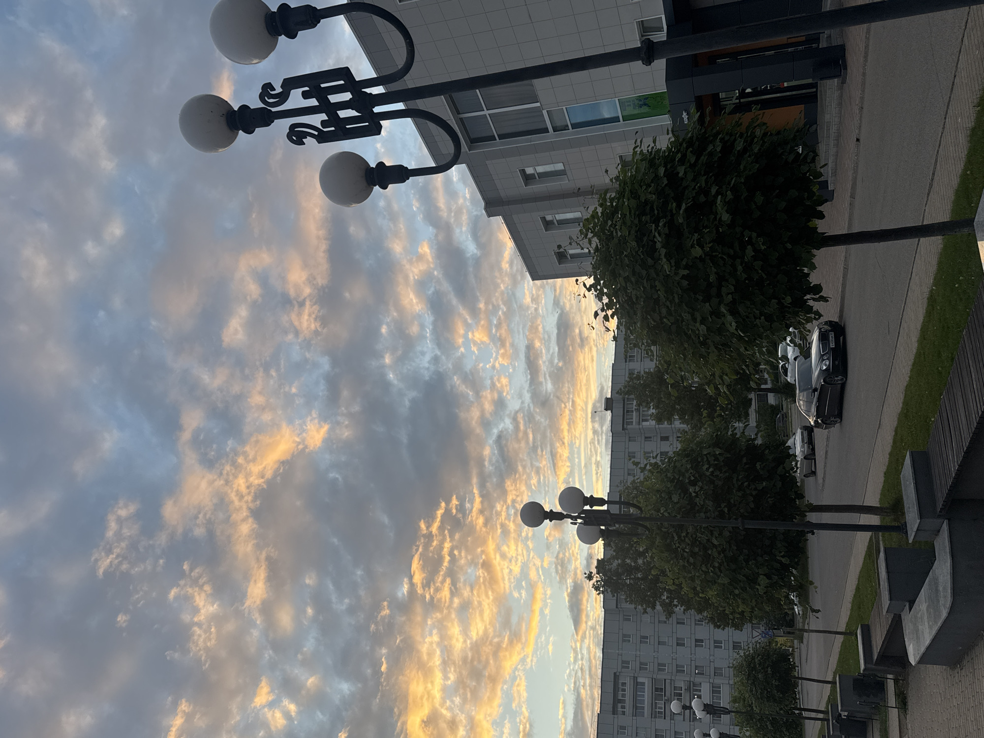 The image depicts a street scene in Chaikovsky, Russia, during twilight. The sky is filled with dramatic clouds illuminated by the setting sun, casting warm orange and pink hues across the sky. In the foreground, a modern lamppost with three spherical lights stands to the right, and another lamppost with multiple lights is visible further down the street to the left. Adjacent to the sidewalk are several concrete benches and planters.
A paved road runs alongside the sidewalk, with two cars visible: a dark-colored sedan, possibly a Mercedes-Benz, and a lighter-colored sedan behind it. Trees line the sidewalk, some appearing neatly trimmed and spherical. Behind the trees and across the street, a large, multi-story residential building with a light-colored facade is prominent, characterized by rows of windows. To the right of the residential building, a modern, grey-tiled building with large windows is partially visible, featuring an entrance with a dark frame and orange accents. The overall impression is of an urban environment transitioning from day to night, with natural beauty in the sky contrasting with the man-made structures.