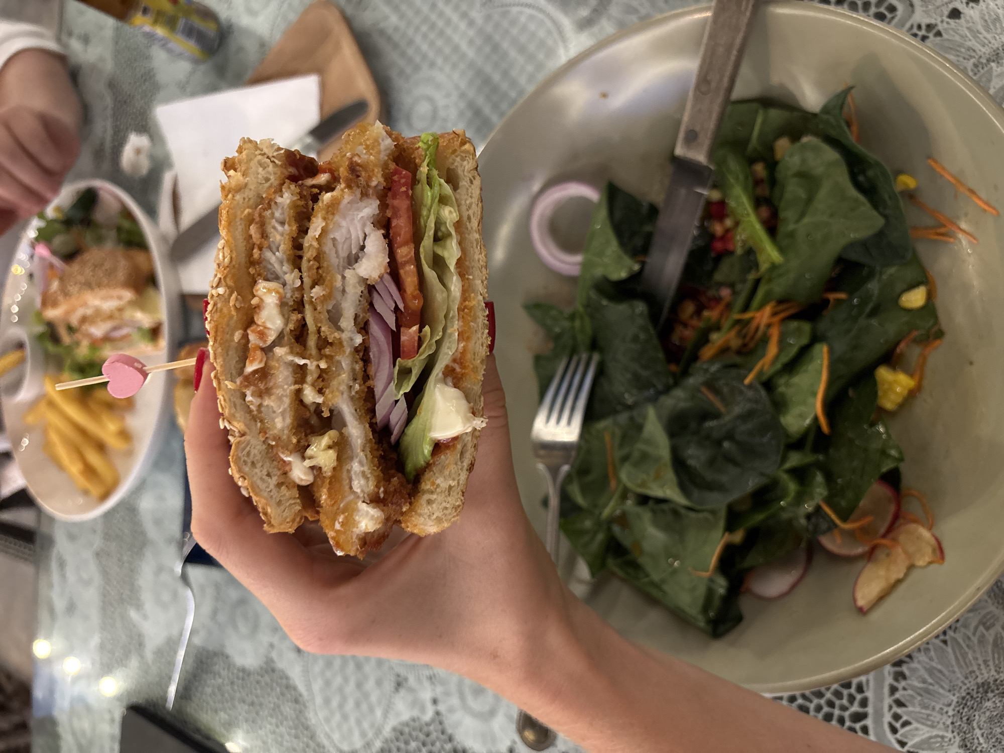 The scene is an indoor restaurant setting. A person is holding a partially eaten fried chicken sandwich that is cut in half, revealing layers of chicken, lettuce, tomato, and red onion. A pink heart-shaped toothpick pierces the sandwich. To the right, there's a bowl of spinach salad with shredded carrots, corn, radishes, and red onion rings. A fork and knife are positioned inside the salad bowl. In the background, there's another plate with what appears to be a similar sandwich and french fries, along with other people and objects on the table. The table is covered with a white, lacy tablecloth. A bottle of condiments and a small wooden cutting board are visible in the background. While the exact location cannot be verified, the image strongly suggests a dining setting with the meal as the primary focus. The phrase Choeng Thale is not recognizable.
