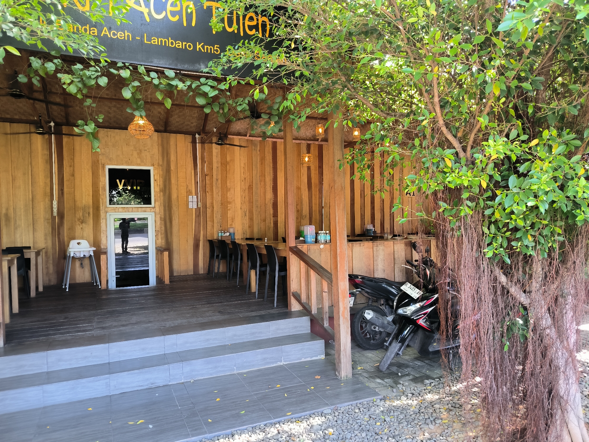 The image shows the exterior of a building in Santan, Indonesia. The structure is primarily constructed with wooden planks. A sign above the entrance reads "Bunda Acen Tulen" and "Inda Aceh - Lambaro Km5". The entrance features three steps leading to a wooden deck. Inside, there are tables and chairs arranged. A high chair is visible near the entrance. A motorcycle is parked to the right of the building, partially obscured by a tree with hanging roots. The area around the base of the steps is covered in small pebbles.