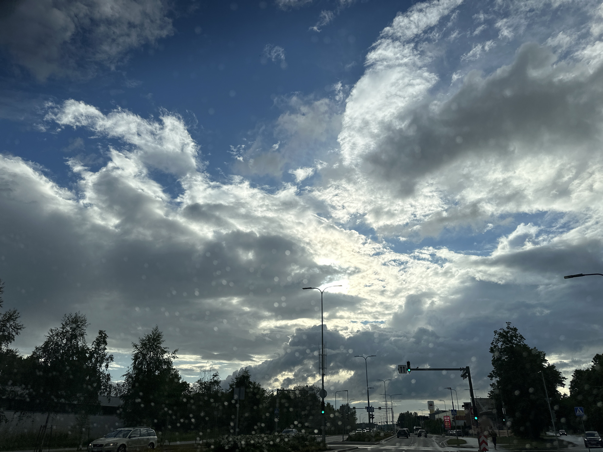 The image shows a street scene in Tallinn, Estonia, on a cloudy day. The sky is filled with dramatic, heavy clouds, with patches of blue sky visible between them. Sunlight is breaking through in some areas, creating bright highlights. There are several cars on the road, and a few trees line the street. Traffic lights are visible, with one showing a green signal. The image appears to have been taken from inside a vehicle, as there are circular water droplets on the windshield.