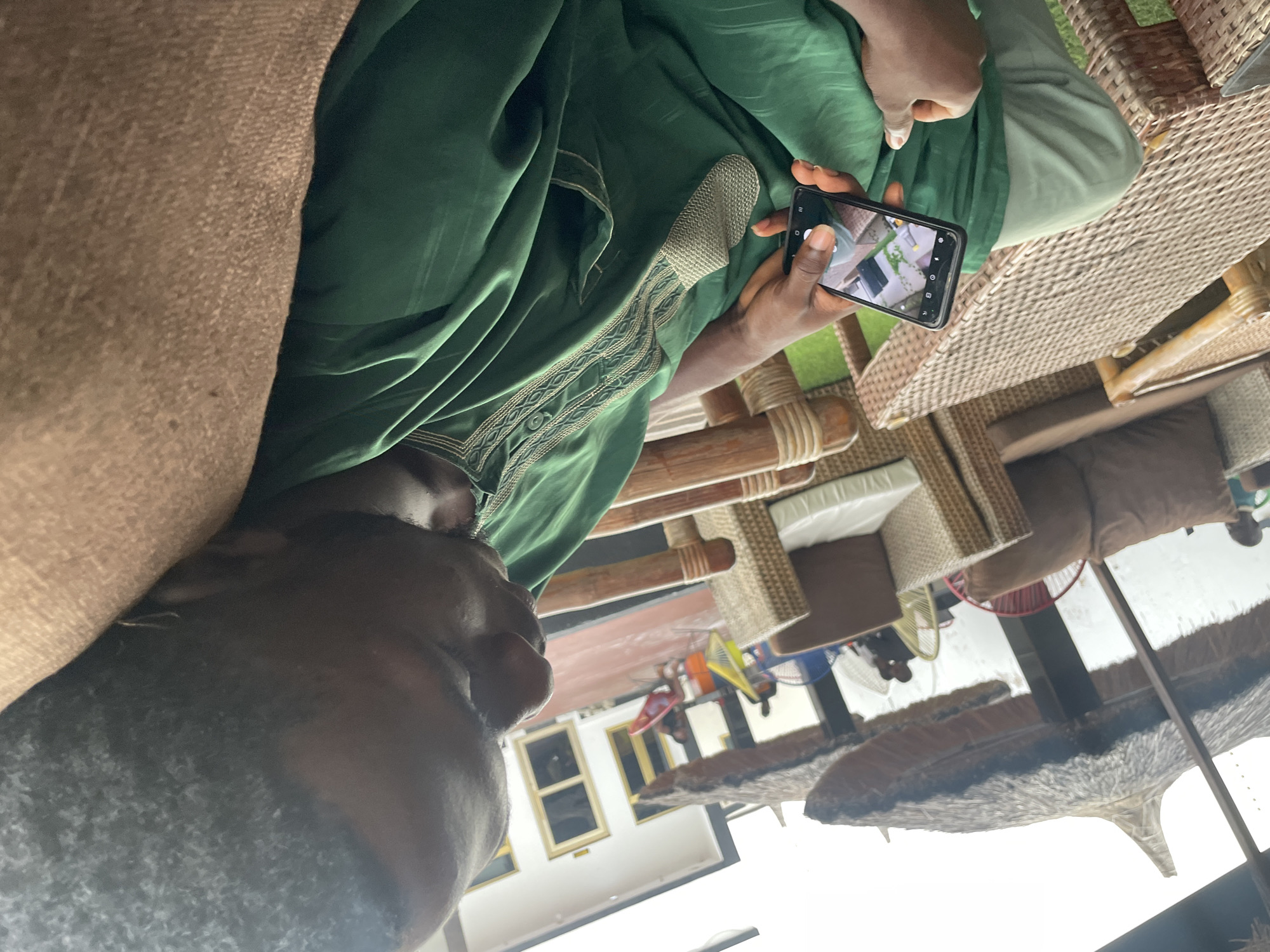The image shows a man in a green traditional Nigerian outfit sitting on a wicker chair and looking at his phone. He is outdoors, possibly at a restaurant or resort, with thatched umbrellas and other wicker furniture visible in the background. The man's face is partially obscured as he focuses on the screen. The environment appears to be a relaxed outdoor seating area with a building and other patrons in the distance. It is not possible to confirm the location as Jauro Yinu, Nigeria, based on the visual information provided.