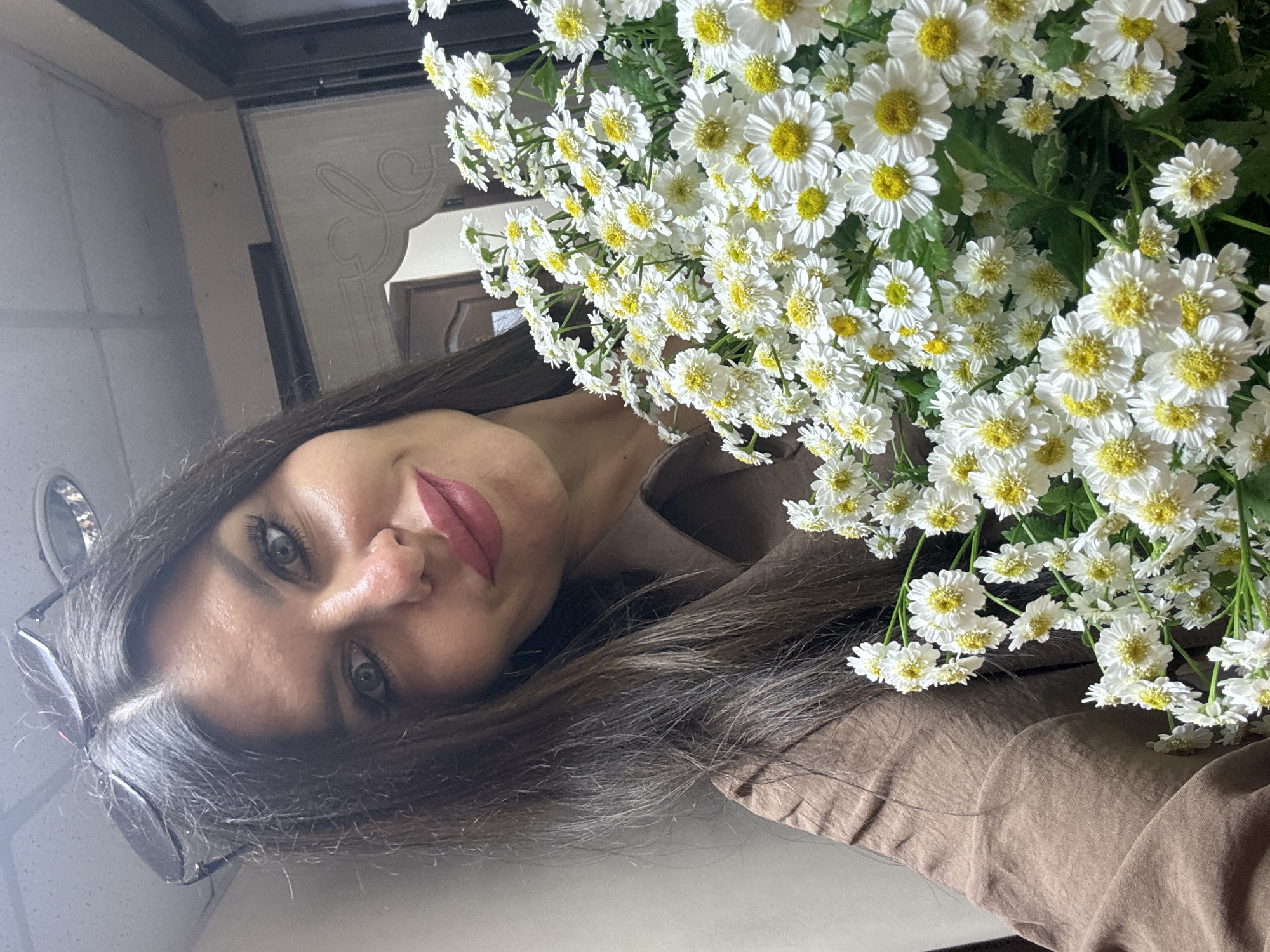 The image shows a young woman holding a bouquet of white daisies with yellow centers. She is looking towards the camera with a soft, pleasant expression. Her long, dark hair is down, and she is wearing a brown top. A pair of sunglasses is perched on her head. The background is out of focus, but appears to be indoors, possibly a room with reflective surfaces and some decor. The lighting suggests it could be daytime. The scene appears to be a candid moment, with the woman and flowers being the central focus. There is no visible text in the image.