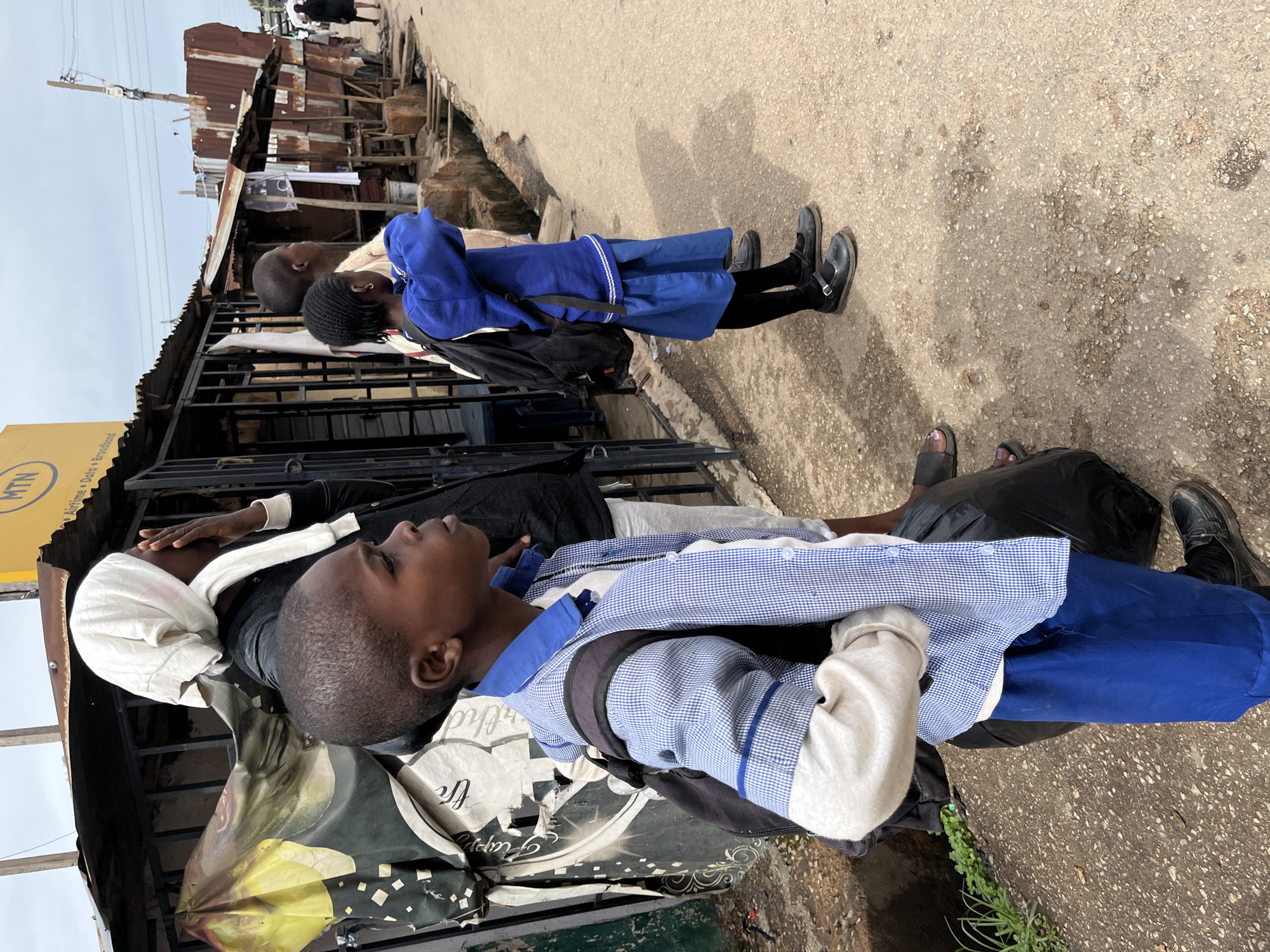 The image captures a street scene in Jos, Nigeria, showing several individuals, primarily children in school uniforms, along a paved road lined with makeshift structures.
In the foreground, a young boy is visible from the chest up, wearing a blue and white checkered uniform shirt over a light-colored long-sleeved shirt, and blue trousers. He carries a dark backpack and looks towards the left side of the frame. Beside him, partially obscured, is an adult or older youth wearing a dark top and a white head covering or cloth, also looking left. Behind these two, another child in a similar blue and white uniform and a dark backpack stands further back, facing right. Further into the background, a child in a full blue uniform, including a blue sweater, stands facing away from the camera. Black school shoes are visible on the feet of the children in uniform. To the far right, the feet of another person wearing black sandals are visible on the pavement.
The setting is an outdoor street, likely bustling, with a rough concrete or asphalt surface. To the left, a series of ramshackle buildings and market stalls constructed from corrugated metal sheets and other materials line the street, suggesting a commercial area. A yellow sign bearing the text "MTN" and "Airline Data & Broadband" is partially visible above these structures. Hanging from one of the structures is a banner or cloth, with the words "Happy Birthday" partially discernible. Some sparse greenery is seen near the bottom right of the frame.
The activity appears to be people standing or waiting on the street, possibly commuting to or from school, or simply pausing during their daily routines. The time of day seems to be daytime, with a bright, possibly overcast sky, indicating ambient light without harsh shadows. The overall atmosphere depicts everyday life in an urban or semi-urban environment. No strong emotions are clearly conveyed by the subjects.