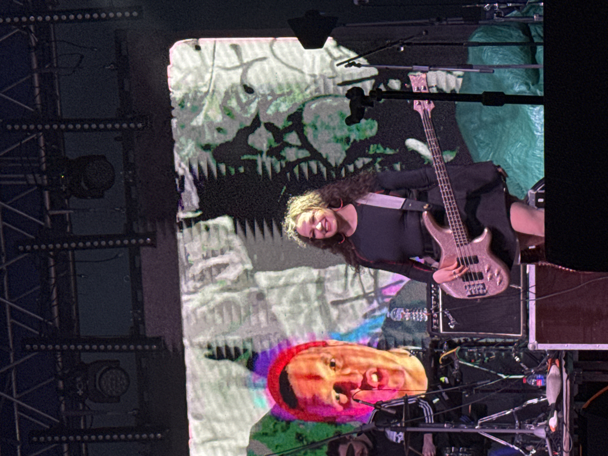 The image depicts a live musical performance on a stage in Большая Кивара, Russia. A female musician, positioned slightly right of center, plays a metallic pink-gold electric bass guitar. She has long, curly brown hair and wears a black long-sleeved top, a black skirt, a light-colored shoulder strap for her bass, and a prominent red hoop earring. She is smiling towards the viewer, with her left hand on the fretboard and her right hand near the pickups.
Behind her, a large LED screen displays abstract digital patterns in green, white, purple, and pink, with visible horizontal pixelation. A large, red-hued projection of a man's face with an open mouth and a red beanie/hat is prominently featured on the left side of the screen.
Overhead, a metal truss structure supports multiple dark, circular LED stage lights. Vertical strips of small, round lights are attached to structural elements on either side of the main screen. To the far left, partially obscured by shadows, another male musician is visible, wearing a black shirt with white stripes on the sleeves, behind what appears to be a drum kit or similar percussion setup.
On the stage floor, a black Fender amplifier cabinet is positioned behind the bass player. Various microphone stands, cables, and other stage equipment are visible, including a small water bottle with a blue cap on a table or case to the left. A large, dark green tarp or covered object rests on the stage floor behind the main performer. The immediate foreground is dark, indicating the edge of the stage.