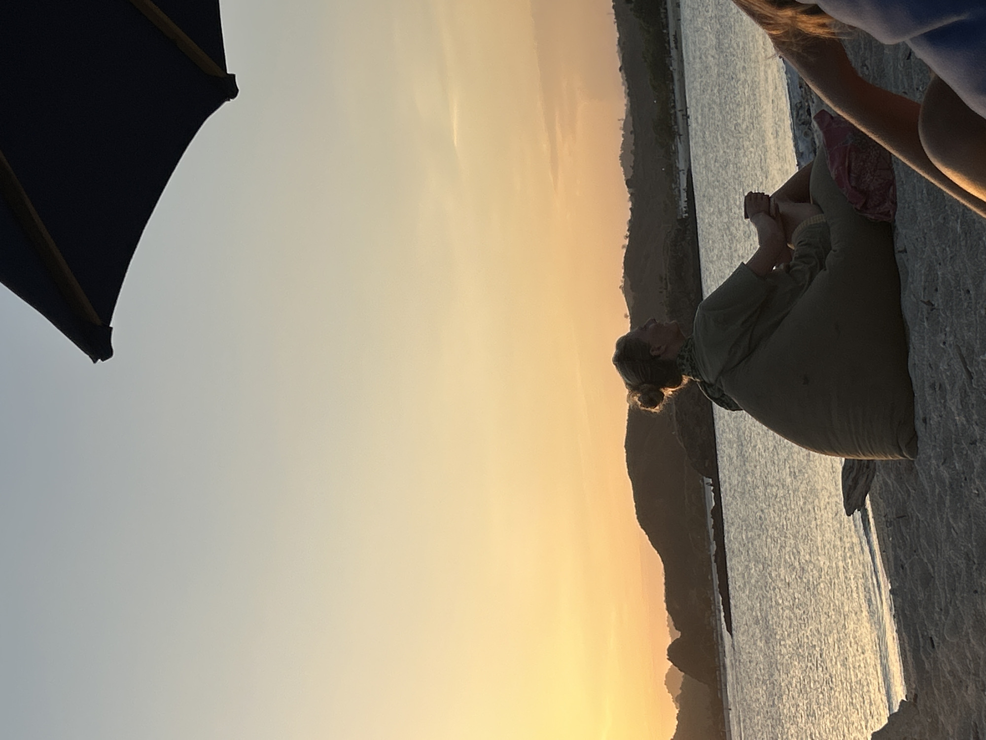 A medium shot captures two individuals on a sandy beach during sunset. In the foreground, a woman sits on a large, taupe beanbag chair, facing away from the camera and towards the ocean. She is wearing a light green t-shirt and has her hair pulled up in a bun. Her hands are clasped in her lap, and she appears relaxed. To the right of the woman, the arm and shoulder of another person are visible, suggesting they are also seated on the beach.
The background reveals a calm body of water, likely the ocean, reflecting the warm hues of the setting sun. Gentle waves lap the shore. Beyond the water, a silhouette of rolling hills and distant mountains is visible against the orange and pale blue sky. A large, dark umbrella dominates the upper right quadrant of the image, casting a shadow over part of the scene. The beach itself is sandy, with a piece of driftwood lying near the beanbag. The overall atmosphere is serene and peaceful, indicative of a beachside location in Kuta, Indonesia, at dusk.