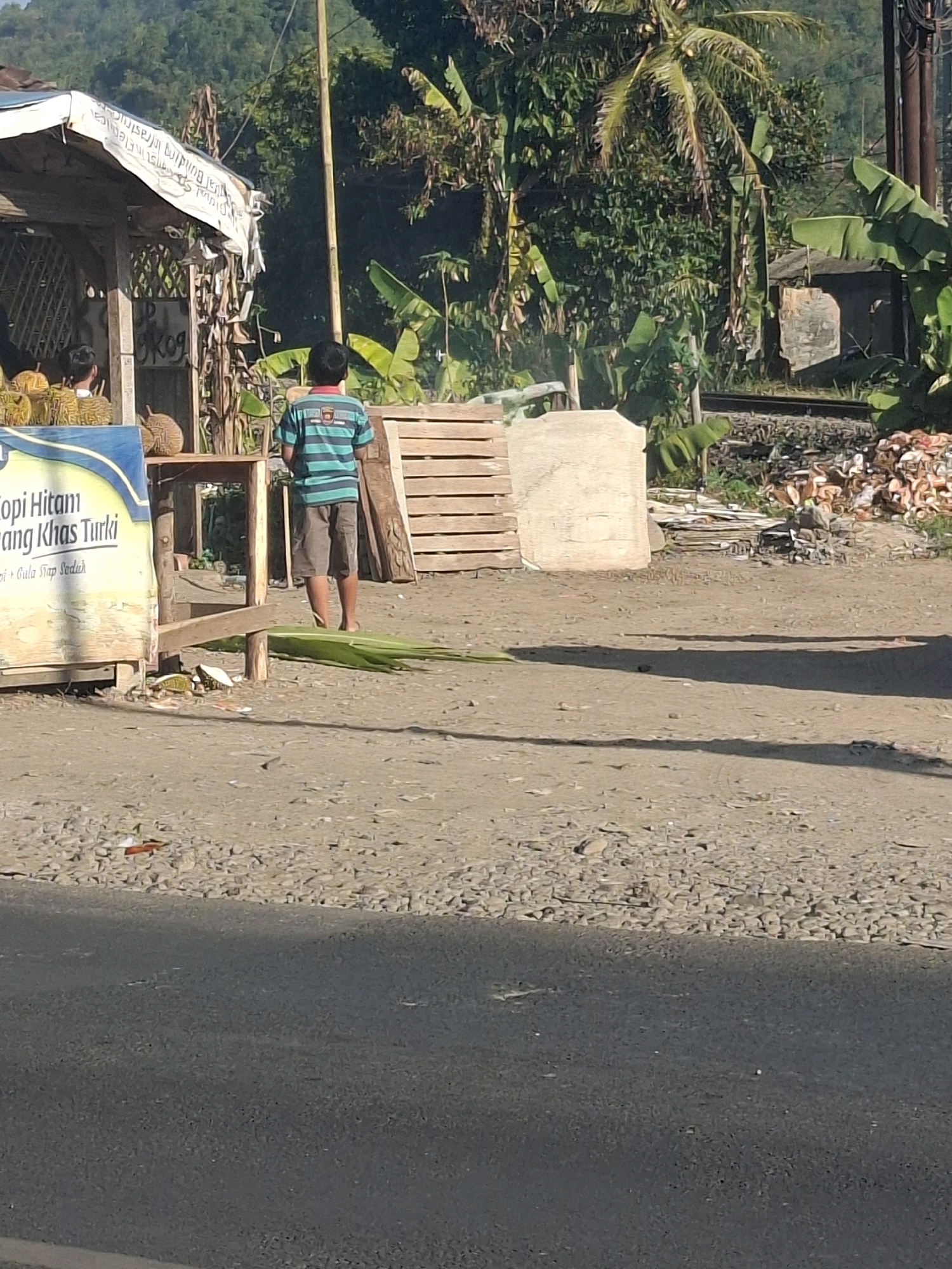 In Lembursawah, Indonesia, a person wearing a horizontally striped t-shirt and shorts stands on a gravelly roadside next to a small stall. The stall displays durian fruit and signage that reads "Kopi Hitam Bang Khas Turki" and "Gula Tiap Seduh." To the right of the stall are stacks of wooden planks and a pile of cut logs. In the background, dense foliage and hills can be seen. The scene appears to be a roadside vendor location.