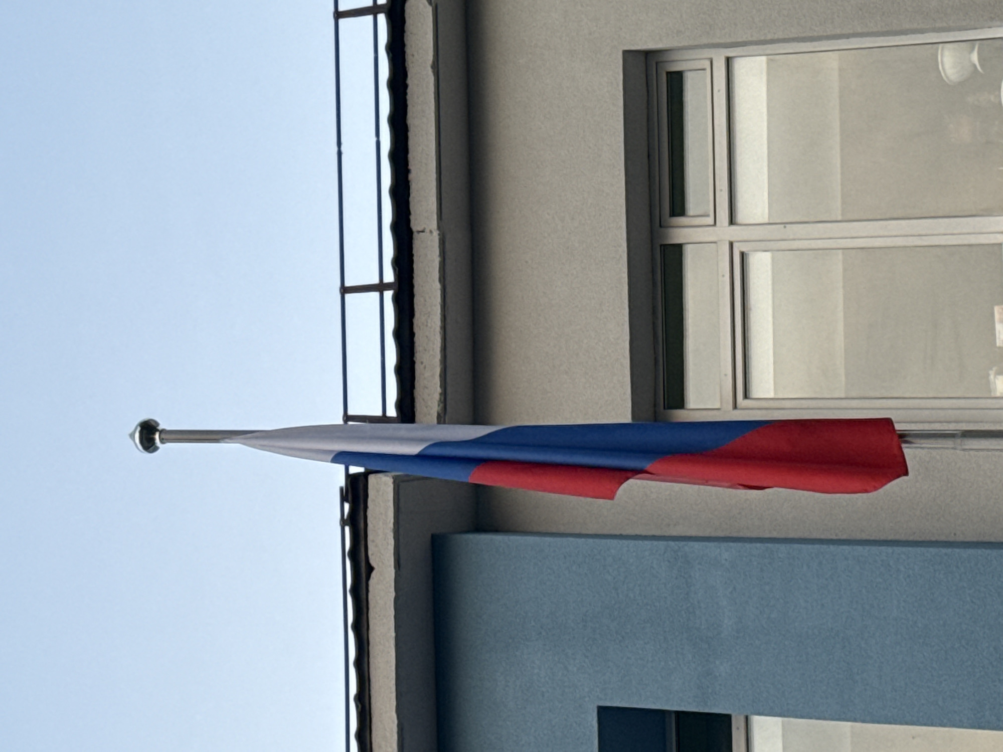 A Russian flag hangs vertically from a flagpole in front of a building. The flag is made of three horizontal stripes: white at the top, blue in the middle, and red at the bottom. The building has two sections: one painted light blue and the other a light gray. Both sections have large windows. A dark railing is visible above the building against a clear blue sky.