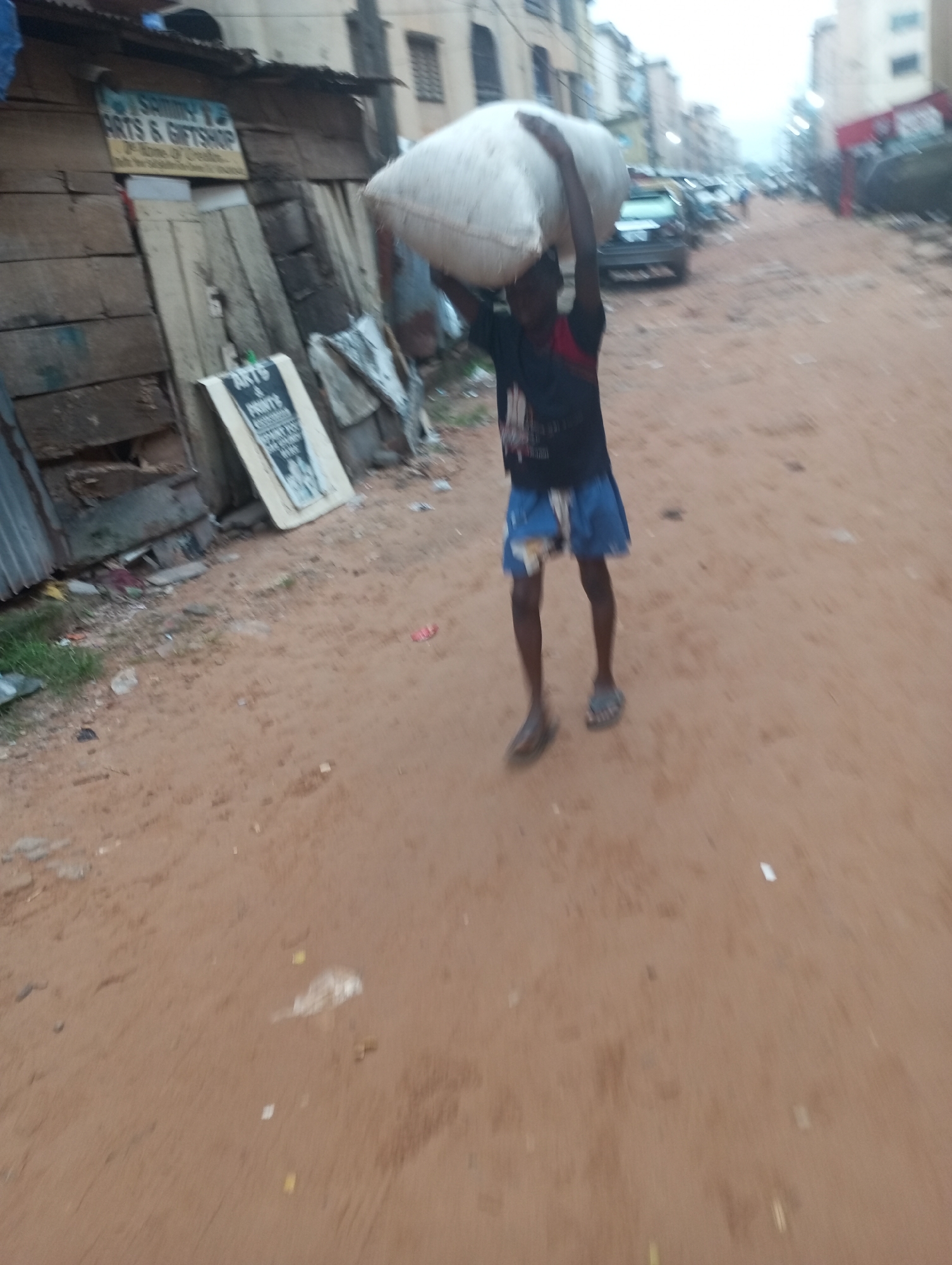 A young person is carrying a large, white bundle of material on their head on a dirt road. The person is wearing a dark t-shirt with a red trim on the sleeves and blue, patterned shorts, as well as sandals. They are walking away from a weathered, wooden structure with a corrugated metal roof. A sign on the structure reads "SAMMY'S ARTS & GIFTSHOP". In the background, cars are parked along the road, and buildings are visible. The ground is reddish-brown dirt with scattered debris. The sky is overcast.