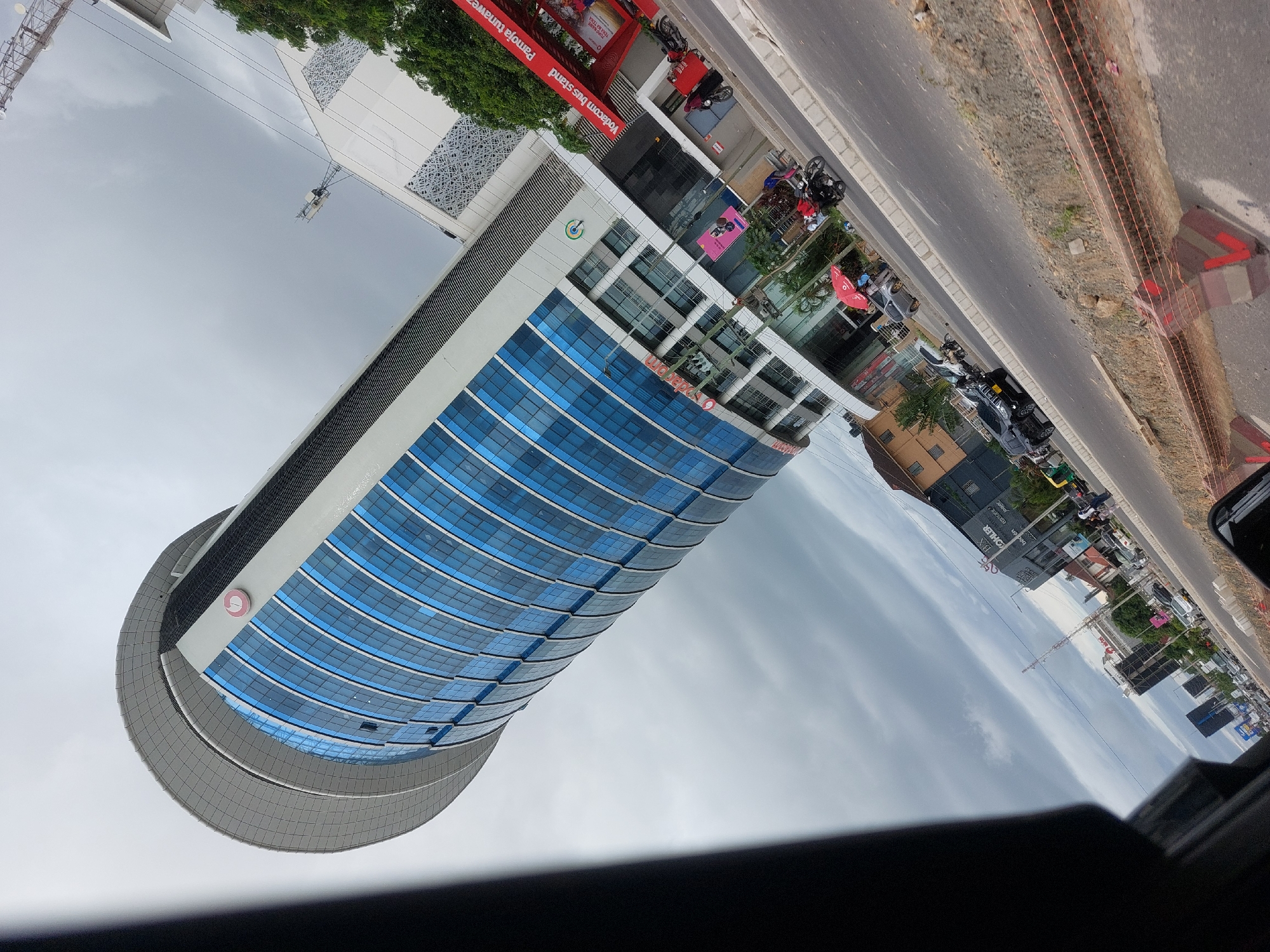 The image shows a tall, modern building with a curved facade of blue glass windows. The building appears to be the Vodacom Tower, located in Dar es-Salaam, Tanzania, identifiable by the Vodacom logo on the building and a Vodacom bus stand nearby. There is a street alongside the building with vehicles and motorcycles parked along it. The sky is overcast with clouds. The photo seems to have been taken from inside a vehicle, evidenced by the dashboard visible at the bottom of the frame. There are no people clearly visible in the image. Construction is evident with orange netting and concrete blocks along the curb.