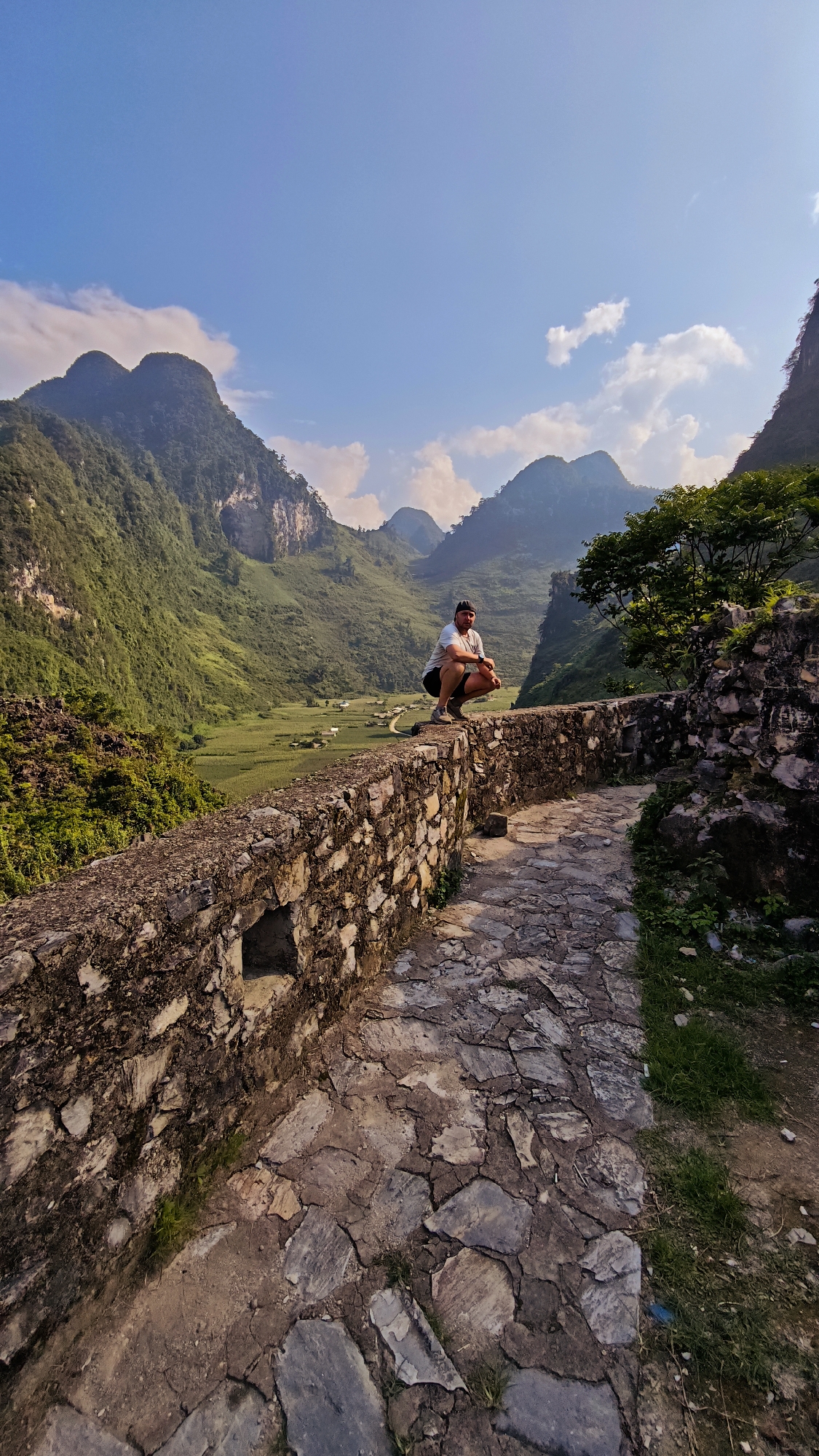A medium-shot captures a man crouching on a stone wall, overlooking a valley in Đường Thượng Commune, Vietnam. The man wears a light-colored T-shirt, dark shorts, and a dark cap. He is positioned on a weathered stone wall that extends into the foreground. A path paved with irregular stones runs adjacent to the wall.
The valley below features lush green vegetation and a cluster of small buildings suggesting a village. Towering mountains form a backdrop, partially obscured by a blue sky dotted with scattered clouds. Vegetation is growing on the wall and adjacent to the path. A small opening is visible in the stonework of the wall.