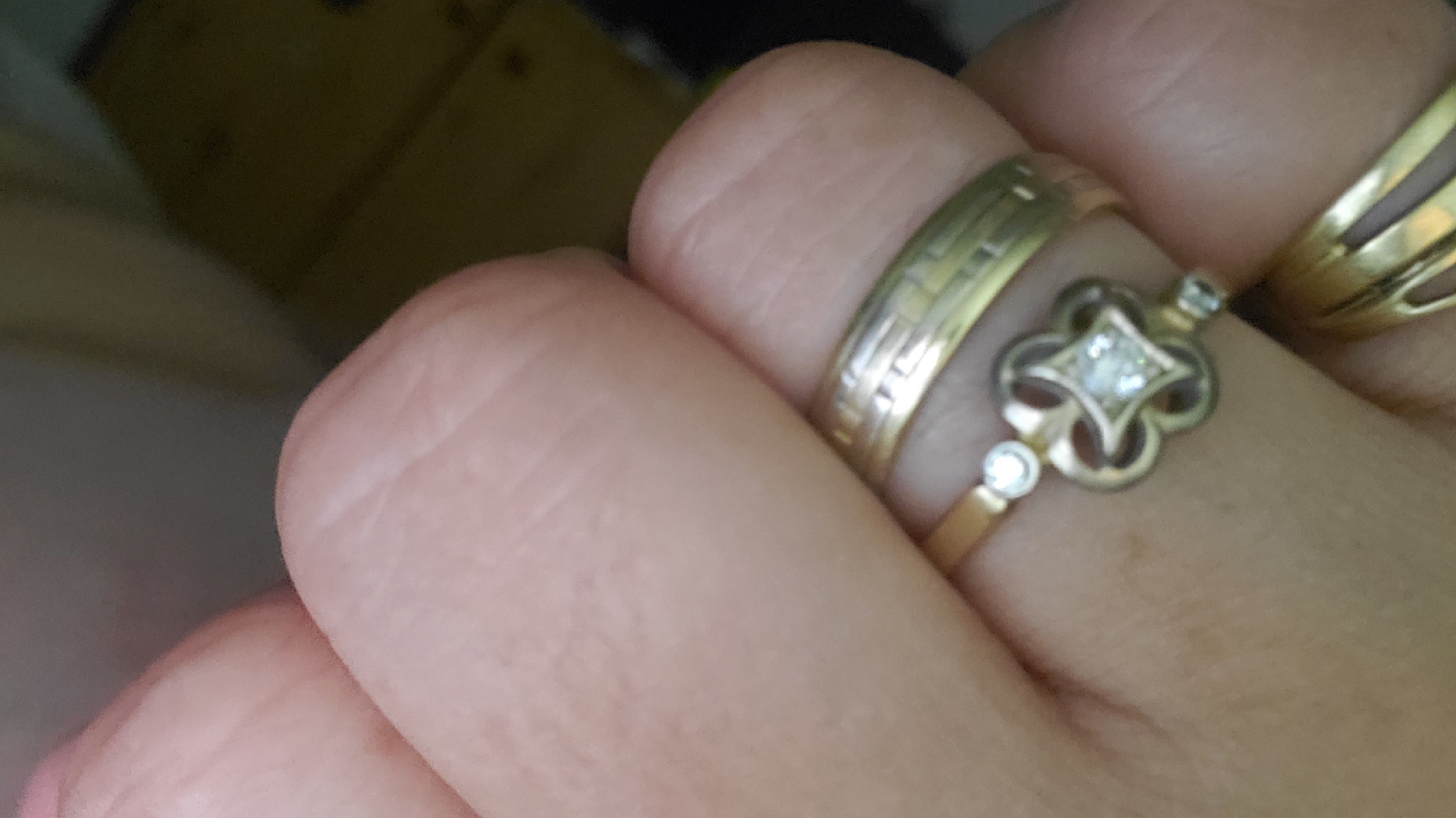 The image shows a close-up of a person's hand wearing three gold rings. The ring closest to the viewer has a band with horizontal lines and two small circular stones on either side. Above this ring is another ring with a clover-like design in the center, featuring a square-shaped diamond, and two smaller circular stones on the band. The third ring, partially visible at the top of the image, is a wider gold band with a split design. The background is out of focus, showing a dark, possibly wooden surface and some indistinct shapes. There are no discernible landmarks or indications of a specific location within the city of Sasbach, Germany, in this image.