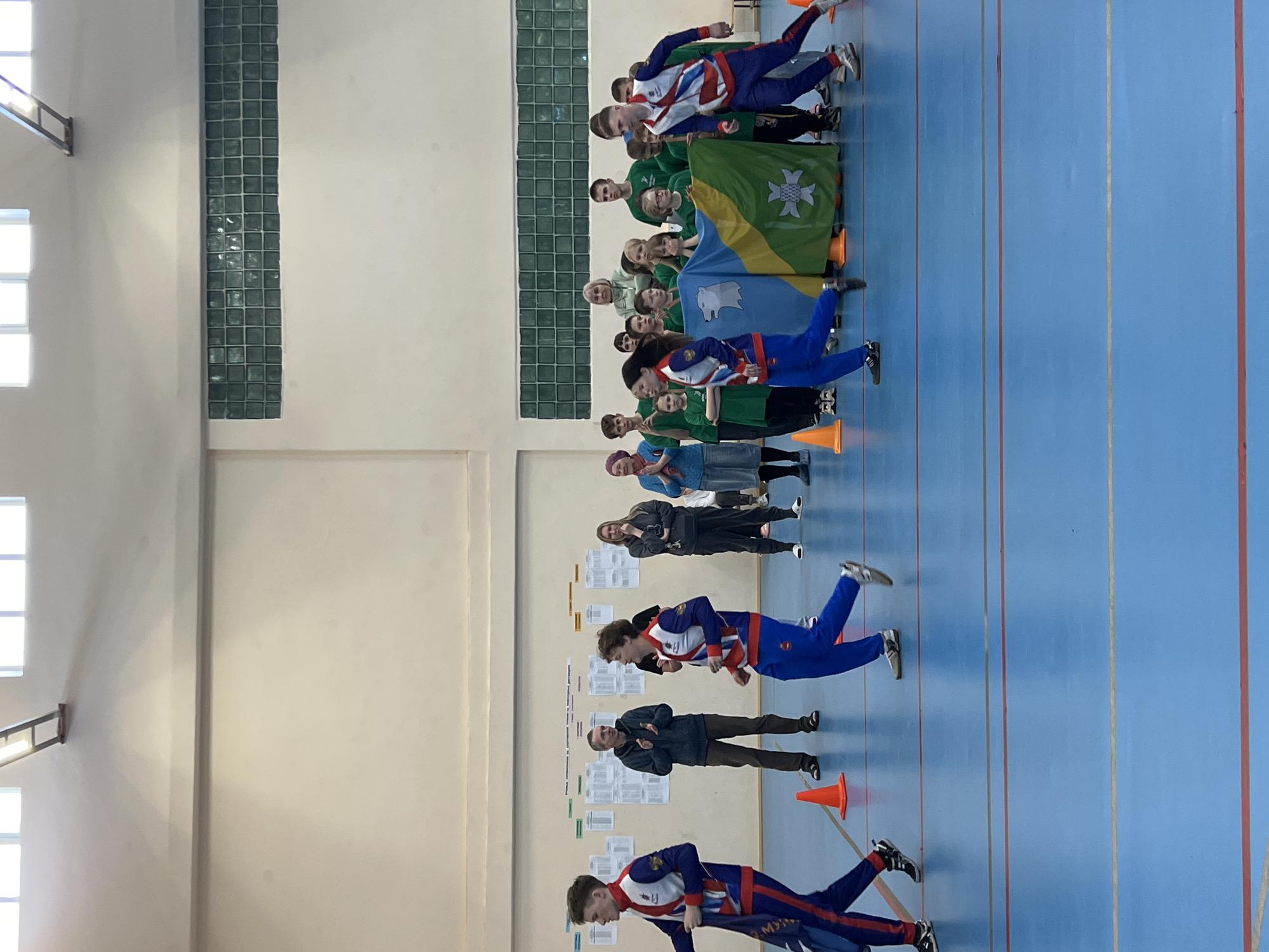 The image depicts a group of people in what appears to be an indoor sports facility, possibly a gymnasium. The location is likely in Berezniki, Russia, based on the flag design that is held by some of the people. There are rows of people, including adults and children. Some are holding what appear to be local or regional flags in shades of green, blue, and yellow. Two individuals are wearing matching red, white, and blue tracksuits. They are performing a crawling exercise along a marked blue floor with lines, past orange cones. Behind them stands an adult male wearing a dark jacket. In the background, there is a wall with an inset of tiled glass bricks, alongside several documents or posters affixed to the wall. The lighting appears to be artificial.