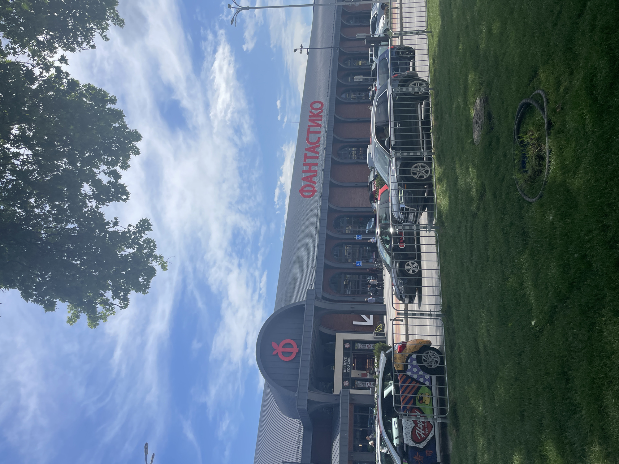 The image depicts the "ФАНТАСТИКО" (Fantastico) supermarket building in Plovdiv, Bulgaria. The facade displays the company logo and name in red lettering. A line of parked cars sits parallel to the building, separated by a metal fence. A grassy area occupies the foreground to the right, including a manhole cover and a circle formed of black electrical cables. A tree canopy partially obscures the sky in the upper left. The sky exhibits a mix of blue and streaky white clouds. The supermarket entrance is visible, marked with an arrow sign.