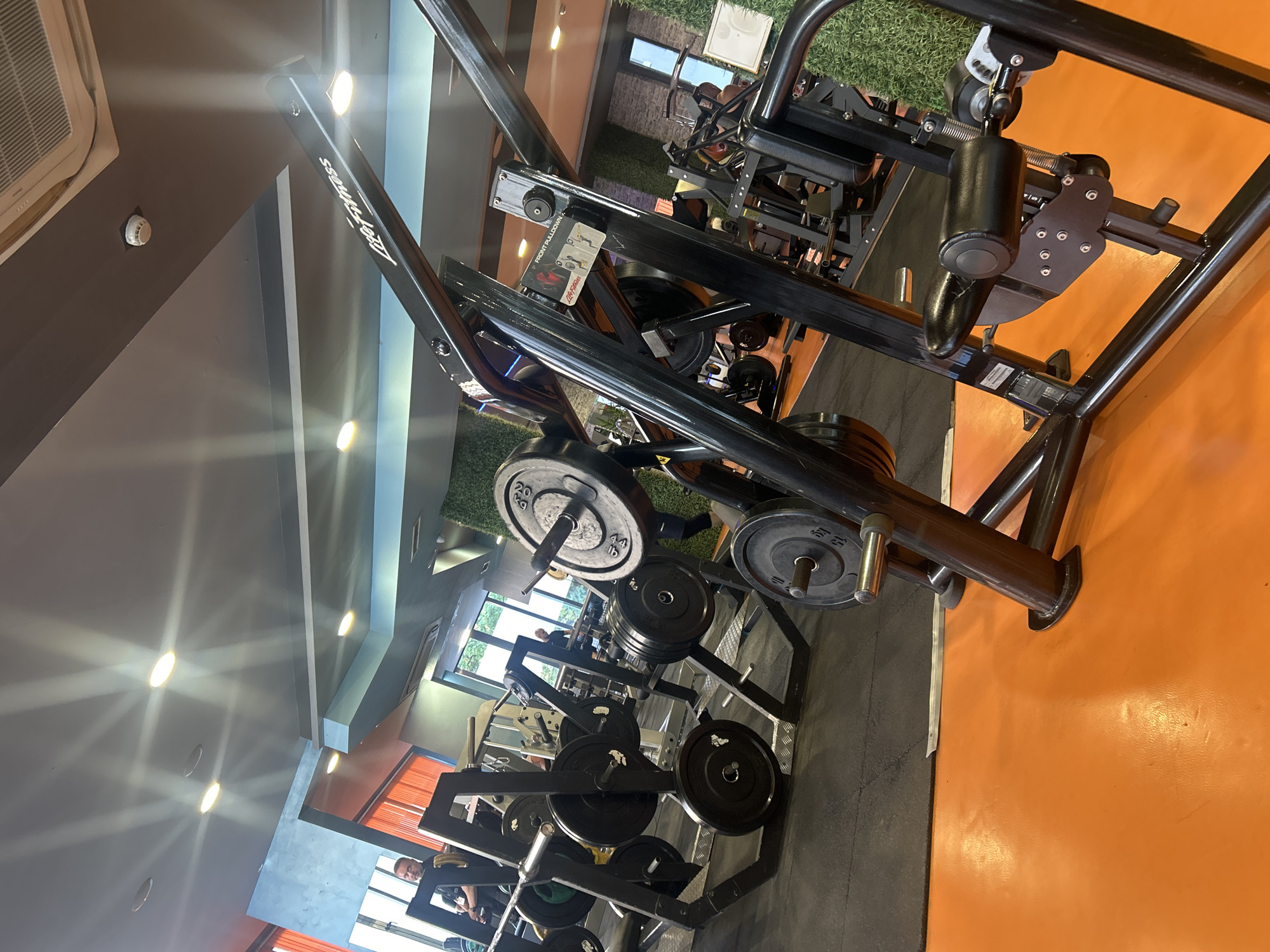 The image depicts the interior of a gym, likely in Sofia, Bulgaria, featuring various weightlifting equipment. The foreground is dominated by a black weight machine with orange flooring visible beneath. The machine has padded black handles and a weight stack. In the midground, several weight racks are loaded with black weight plates, ranging in size and weight, including some marked with "20 KG" and "15 KG". A barbell rests on one of the racks. The background reveals more gym equipment, a man in a dark shirt standing near a machine, and large windows letting in natural light. The walls are painted in shades of blue and gray, with orange accents. Artificial green walls, possibly decorative, are visible in some areas. The ceiling is gray with recessed lighting and an air conditioning unit. The overall impression is of a well-equipped fitness facility.