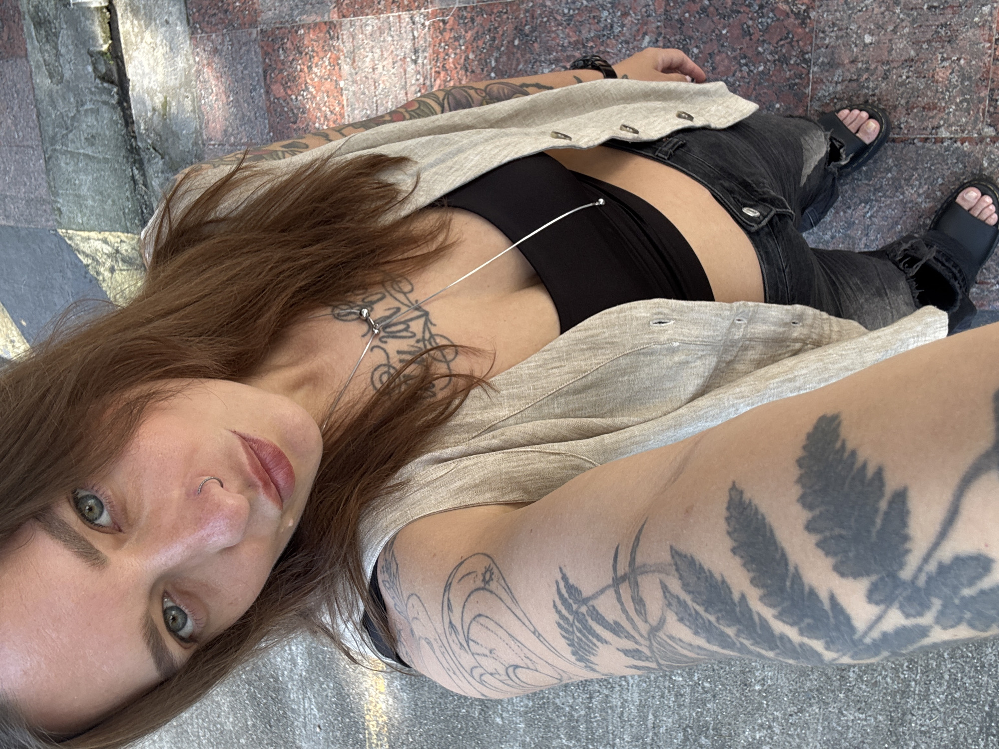 A young adult female is depicted in a selfie taken outdoors in an urban environment, identified as the city of Sochi, Russia. The subject has medium-long reddish-brown hair, green eyes, and a silver hoop piercing in her left nostril. She is wearing a black crop top, a light beige sleeveless vest with button closures, and distressed dark grey jeans with rips. Her footwear consists of black open-toed sandals. A long silver necklace with two spherical elements hangs around her neck. Extensive tattoos are visible: a detailed fern leaf design covers her left forearm, extending towards the upper arm with additional linear and abstract patterns, and her right arm and upper chest display intricate floral or ornamental designs. The ground beneath the subject is composed of two distinct surfaces: light grey concrete or asphalt pavement on the left and reddish-brown polished granite or marble tiles on the right. The scene is illuminated by bright natural light.
