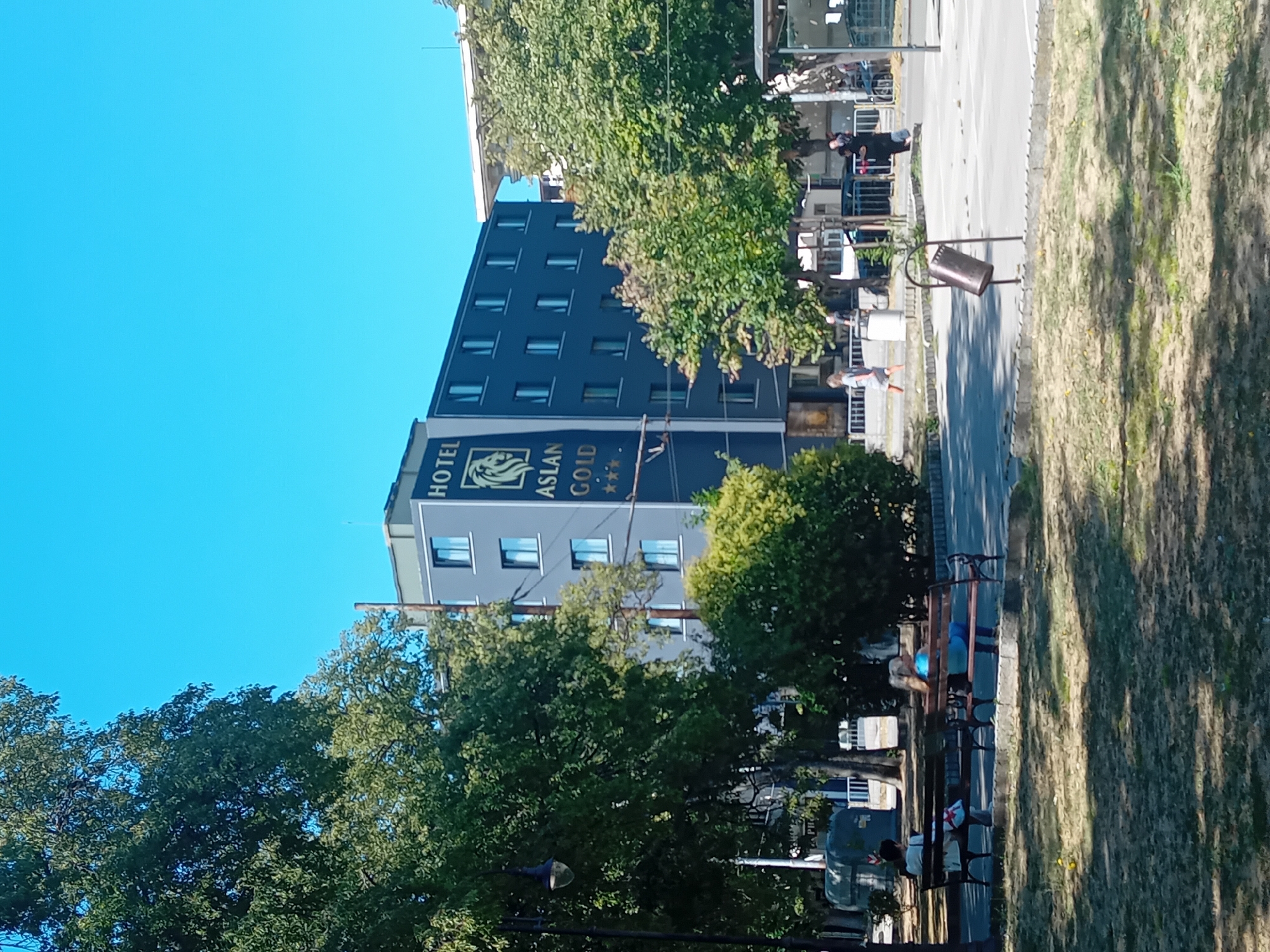 The image depicts a sunny day in Varna, Bulgaria, with the "Hotel Aslan Gold" building prominently featured. The hotel is a multi-story structure with a dark facade and numerous windows. The hotel's name and a logo featuring a lion are visible in gold lettering on its side. In front of the hotel, there is a paved area with a few people walking and standing. Some trees and green spaces are also visible, suggesting a park or a public area adjacent to the hotel. Two individuals are seated on benches in the foreground, facing away from the camera, seemingly enjoying the park. The environment appears to be urban, with a clear blue sky overhead.