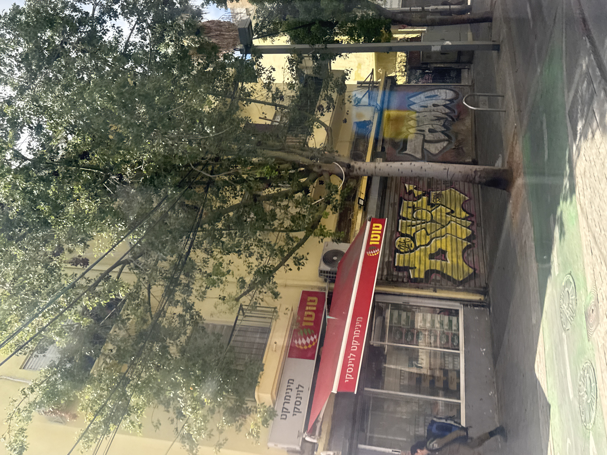 A street scene in Tel-Aviv, Israel, depicts a store, identified by signage in Hebrew reading "מינימרקט לוינסקי" and "טוטו". The store is adjacent to a sidewalk, where a person is walking. The store's facade is adorned with graffiti. A tree is located near the store.