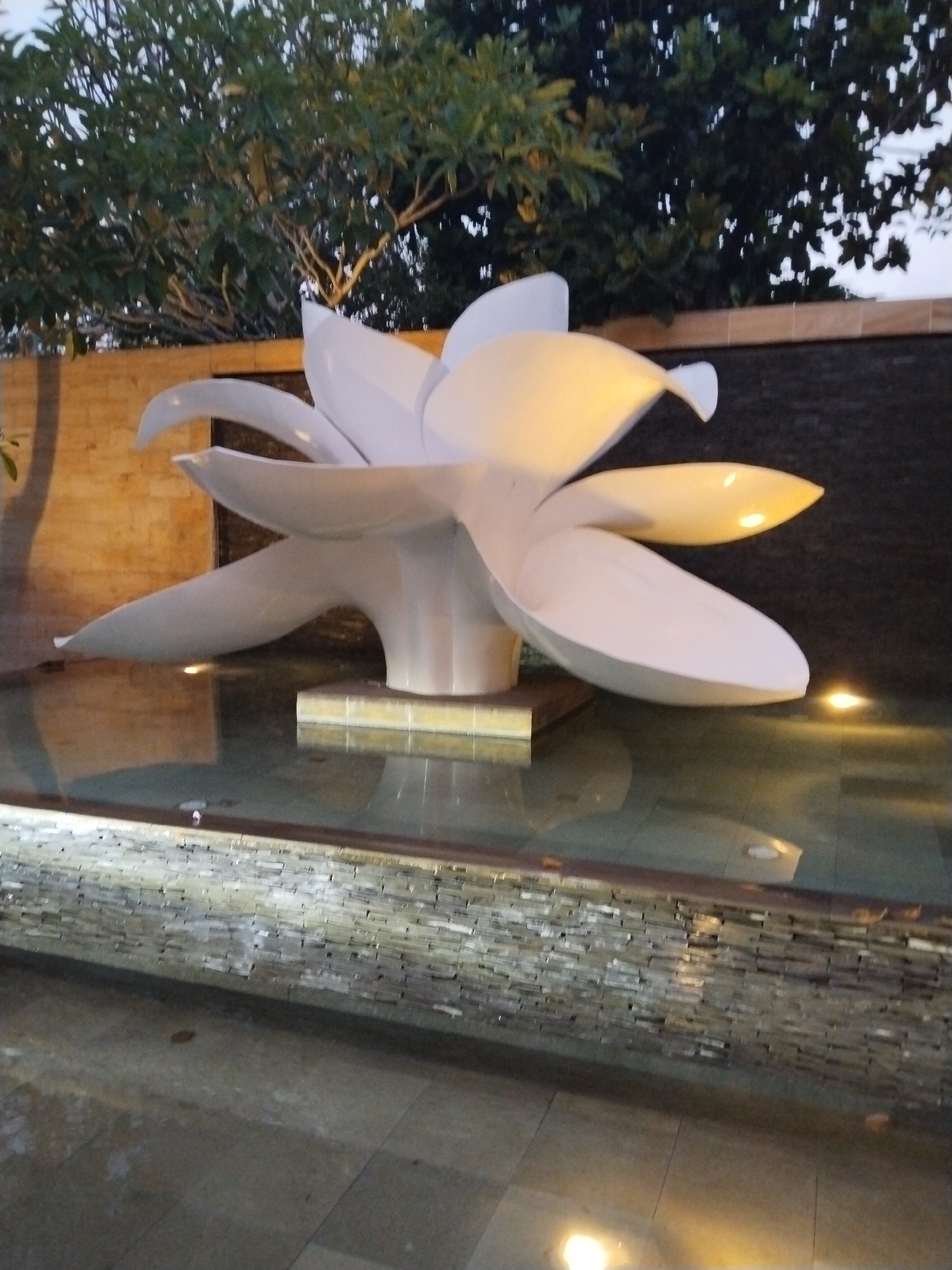 The image depicts a white lotus flower sculpture in a shallow pool of water, likely part of an outdoor decorative water feature. The sculpture is prominently illuminated. The pool is surrounded by a brick or stone border with a textured wall behind it. Trees and foliage are visible in the background. The ground in the foreground is tiled. This appears to be a tranquil public space in Kuta Selatan, Indonesia, possibly a hotel or resort. No people are visible in the image.