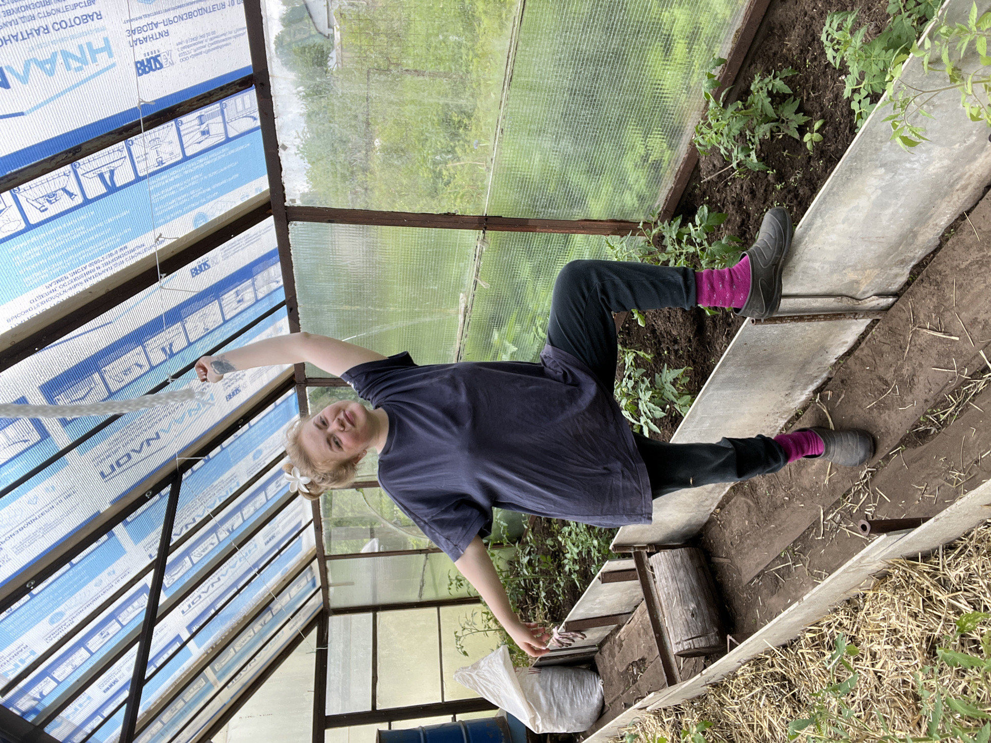 The image captures a woman inside a greenhouse, possibly located in the city of Chaikovsky, Russia, based on text detected on the greenhouse material. The greenhouse's structure is primarily constructed of metal beams and some type of translucent material. The woman is standing diagonally within the frame, facing towards the camera. She is dressed in a dark-colored t-shirt, dark pants, and pink socks, with one foot clad in a dark-colored clog-style shoe. Her hair is pulled back, and she seems to be holding or adjusting a string or rope attached to the greenhouse structure above. The greenhouse contains garden beds with planted greenery, and there are several other objects, including a bucket and bags, within the space. The exterior view through the greenhouse walls reveals greenery.