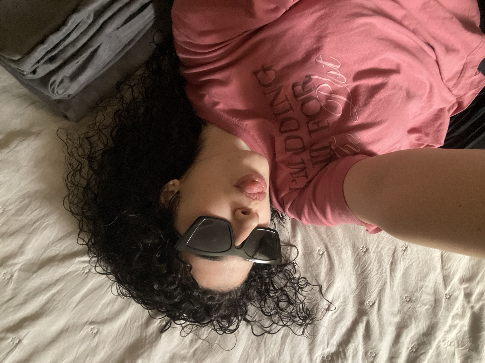 A woman is lying on a white bed, wearing a pink t-shirt and black sunglasses. Her curly dark hair is spread out on the bed. The bed has a white, embroidered duvet cover. There is a folded gray blanket on the bed. The woman is sticking her tongue out. This image does not provide information regarding a location in Sofia, Bulgaria.