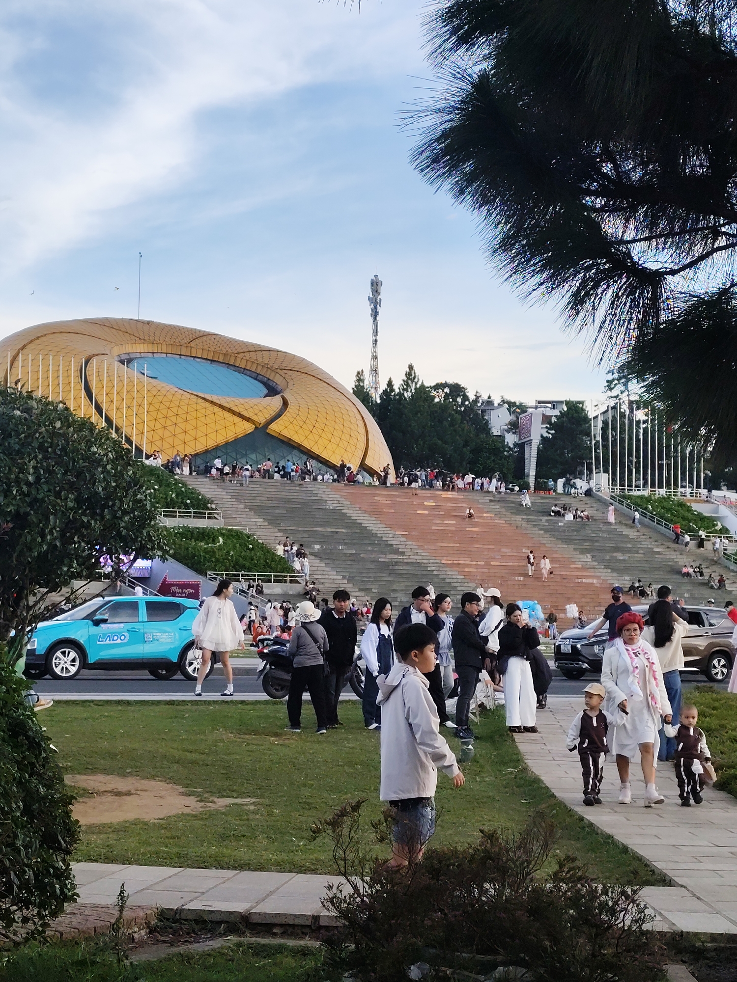 In an outdoor urban setting, a group of people are gathered near a large, modern, gold-domed building with tiered stone steps leading up to it. The sky is partly cloudy. A bright blue car with "LADO" branding is parked on the left side of the image, in front of a grassy area. Several individuals and families are seen walking on the steps and the paved walkway. Some are seated on the steps, while others are walking towards the building or away from it. A man in a light-colored jacket and shorts is walking on the grass towards the foreground, while a woman in a white dress and two young children in matching brown and white outfits are walking on the paved path in the foreground. Other people are visible in the midground, including a woman with red hair wearing a white jacket and scarf. Behind the building, a tall antenna tower is visible. The overall scene suggests a public gathering or event at a cultural or recreational venue.