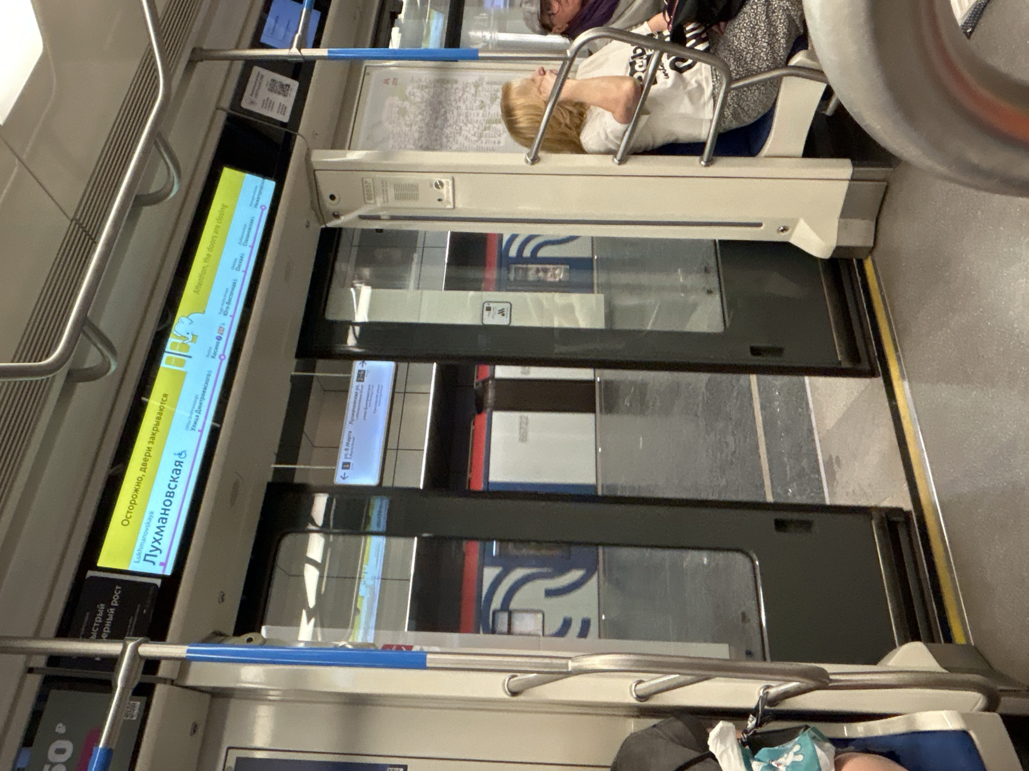 The image shows the interior of a subway train. A digital display above the doors shows "Осторожно, двери закрываются" (Caution, doors are closing) in Russian, and "Attention, the doors are closing" in English, with the station name "Лухмановская&" (Lukhmanovskaya) visible. Below the display, a map of the Moscow Metro system is partially visible. Two passengers are seated on the right side of the train, with one woman in a white t-shirt and patterned pants visible from the chest up, and another person wearing a cap. The train doors are open, revealing a subway platform outside. The platform is wet and reflective, with a train on the opposite track visible. The train car interior is clean and modern, with metal poles for support and beige and white paneling.