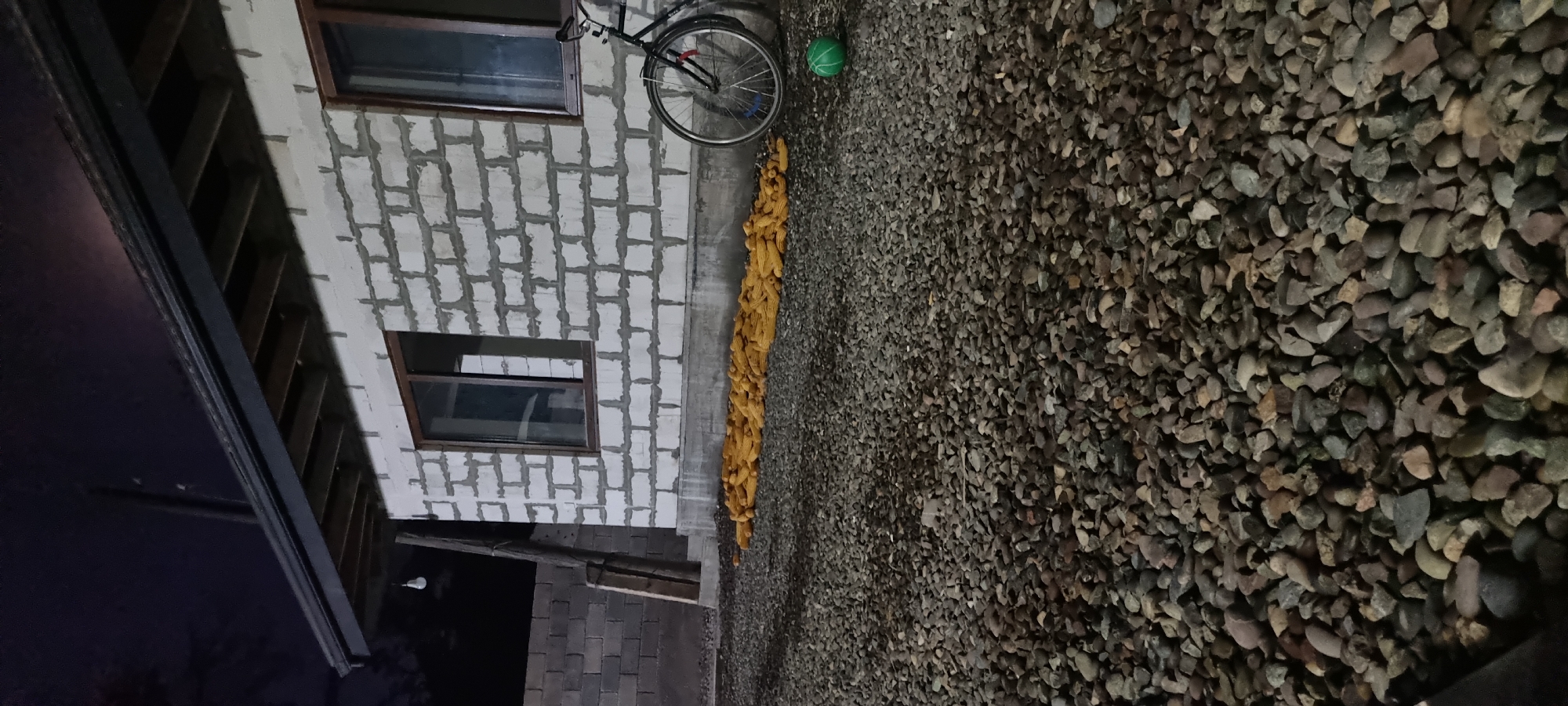The image depicts an outdoor scene at night in Jomi, Tajikistan. A building under construction, with white cinder blocks and visible mortar, forms the backdrop. The building has two windows with dark frames. A black bicycle with a front basket is parked near the right side of the building. In front of the building, a large pile of dried corn cobs is spread out on the gravel ground. To the right of the corn, a small green ball is visible on the gravel. The ground is covered in a layer of small, multi-colored rocks. A partially visible brick wall is present to the left of the building. A wooden post with some wire is leaning against the left side of the building, near the brick wall. The sky is dark, suggesting nighttime, and a light bulb hangs from the eaves of the building.